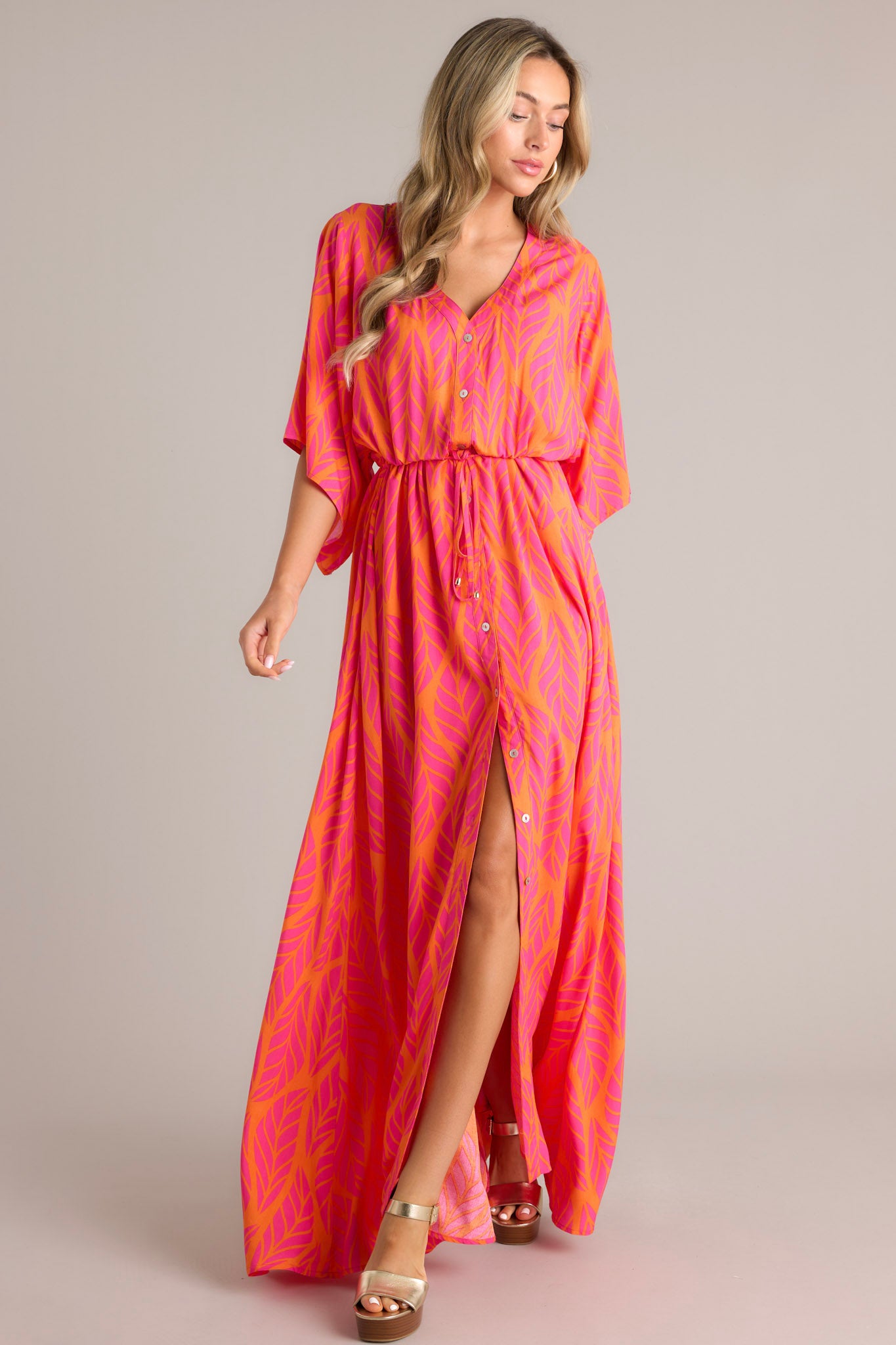 Full body view of this hot pink maxi dress that features a v-neckline, a functional button front, a self-tie drawstring waist, a flowing silhouette, and flowing half sleeves.