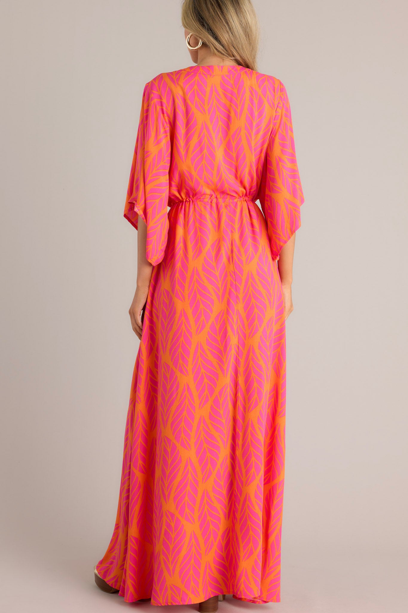 Back view of this hot pink maxi dress that features a v-neckline, a functional button front, a self-tie drawstring waist, a flowing silhouette, and flowing half sleeves.