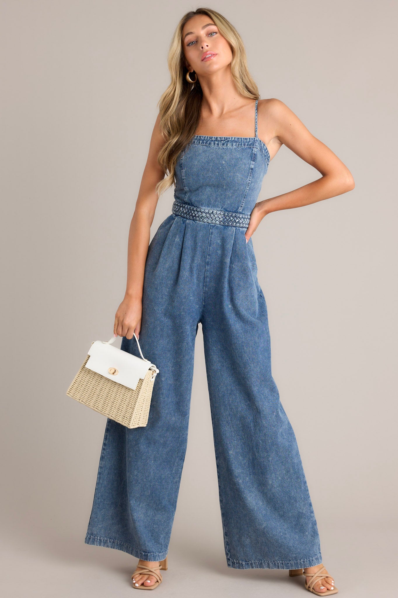 Full body view of this dark chambray jumpsuit that features a square neckline, thin adjustable straps, a discrete back zipper, a thick waistband, subtle pleats, functional hip pockets, and a wide leg.