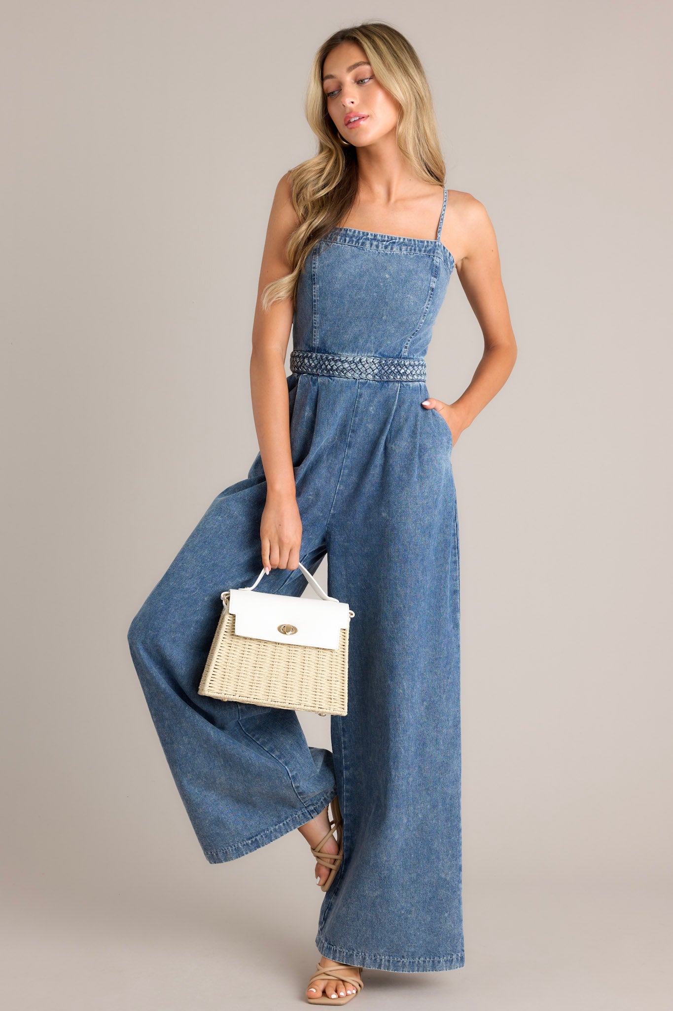 Angled full body view of this dark chambray jumpsuit that features a square neckline, thin adjustable straps, a discrete back zipper, a thick waistband, subtle pleats, functional hip pockets, and a wide leg.