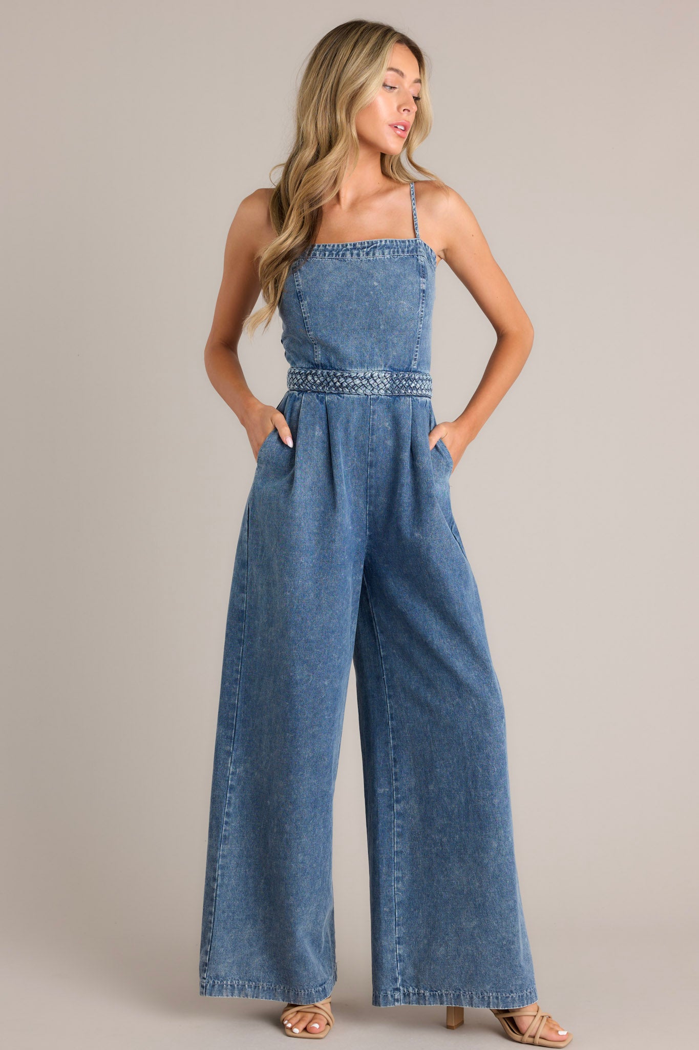 Angled full body view of this dark chambray jumpsuit that features a square neckline, thin adjustable straps, a discrete back zipper, a thick waistband, subtle pleats, functional hip pockets, and a wide leg.