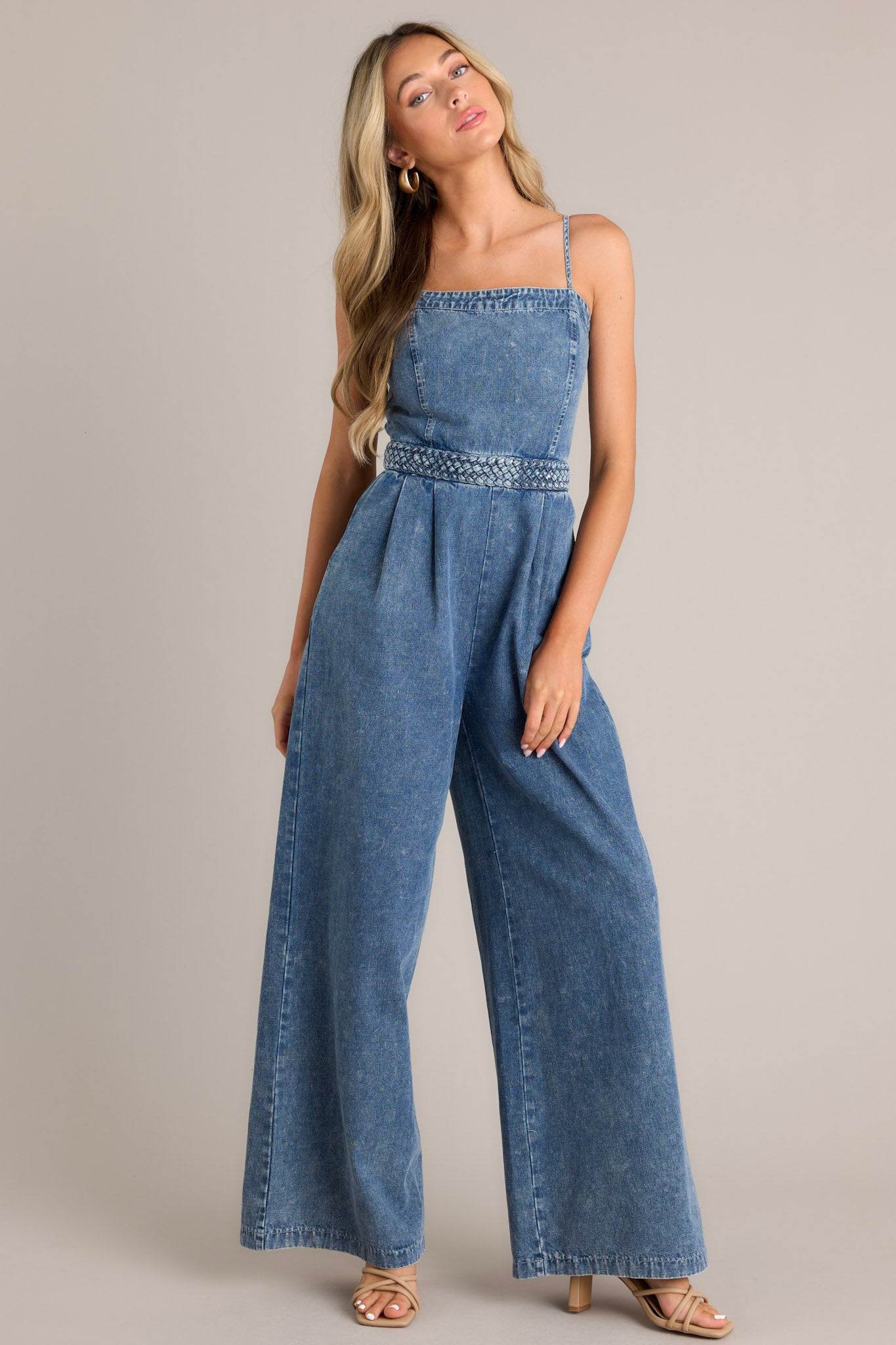 Front view of this dark chambray jumpsuit that features a square neckline, thin adjustable straps, a discrete back zipper, a thick waistband, subtle pleats, functional hip pockets, and a wide leg.