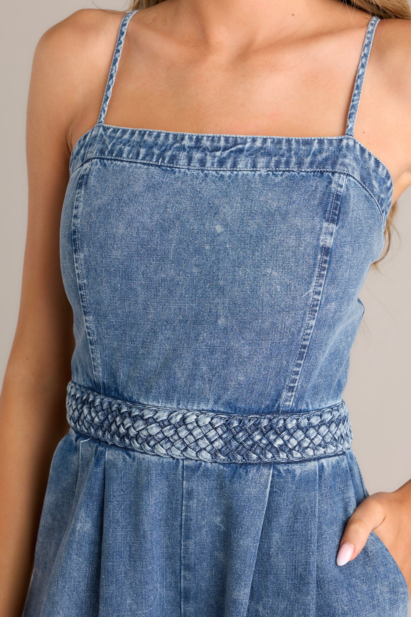 Close up view of this dark chambray jumpsuit that features a square neckline, thin adjustable straps, a discrete back zipper, a thick waistband, subtle pleats, functional hip pockets, and a wide leg.