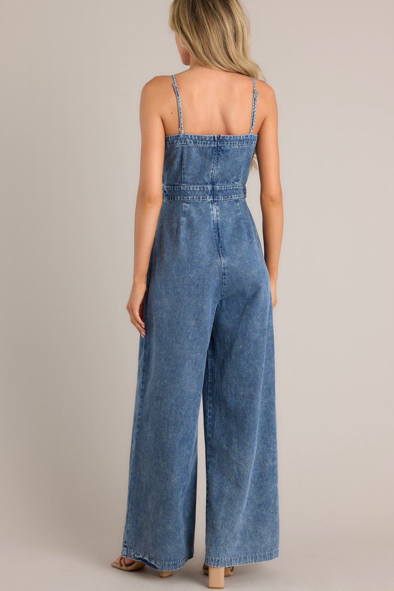 Back view of this dark chambray jumpsuit that features a square neckline, thin adjustable straps, a discrete back zipper, a thick waistband, subtle pleats, functional hip pockets, and a wide leg.