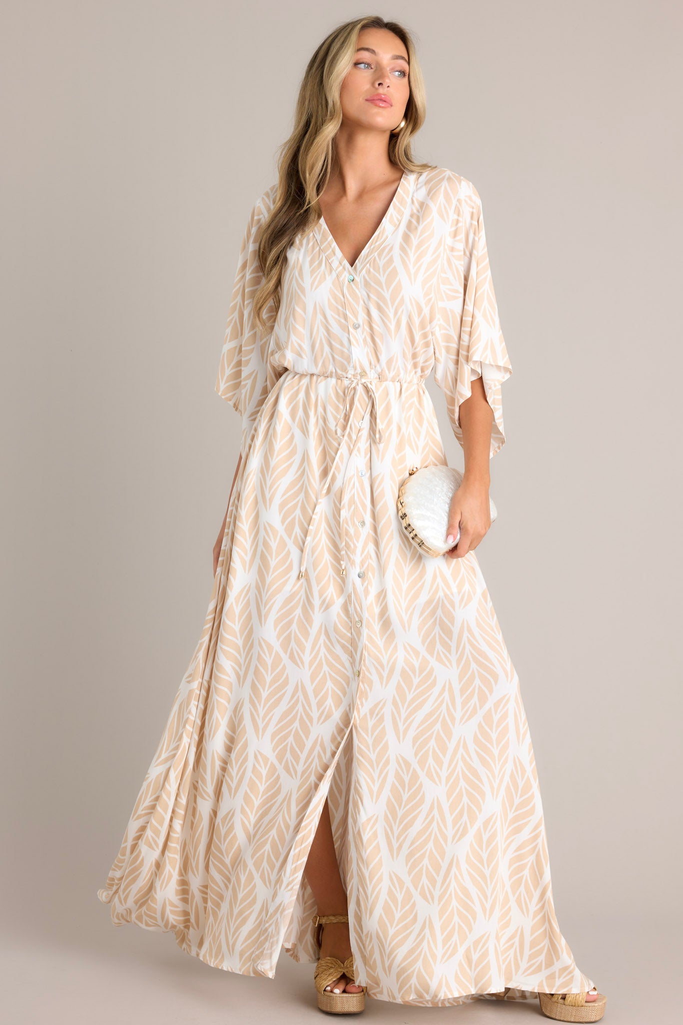 Angled full body  view of this beige maxi dress that features a v-neckline, a functional button front, a self-tie drawstring waist, a flowing silhouette, and flowing half sleeves.