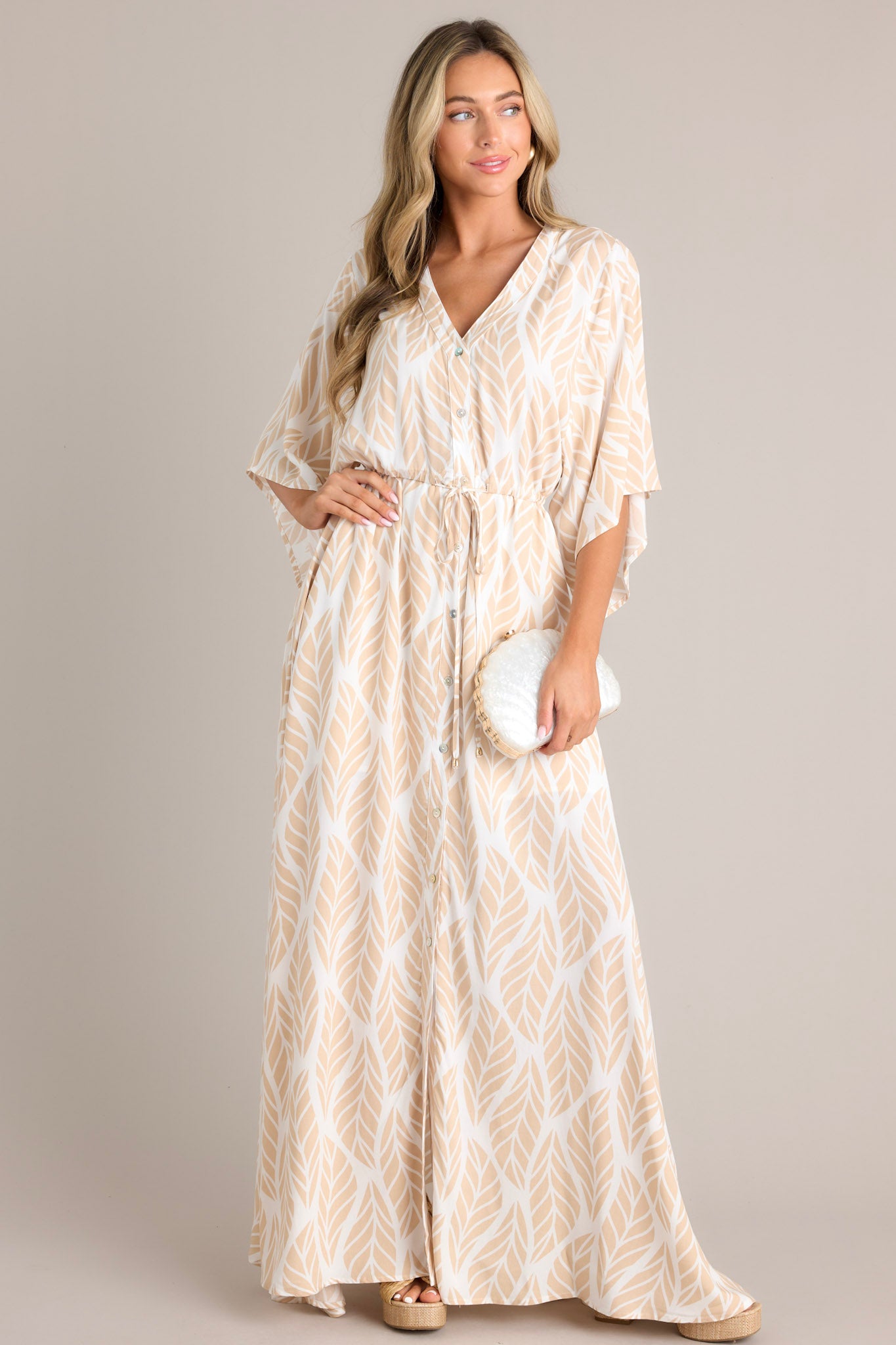 Full body  view of this beige maxi dress that features a v-neckline, a functional button front, a self-tie drawstring waist, a flowing silhouette, and flowing half sleeves.