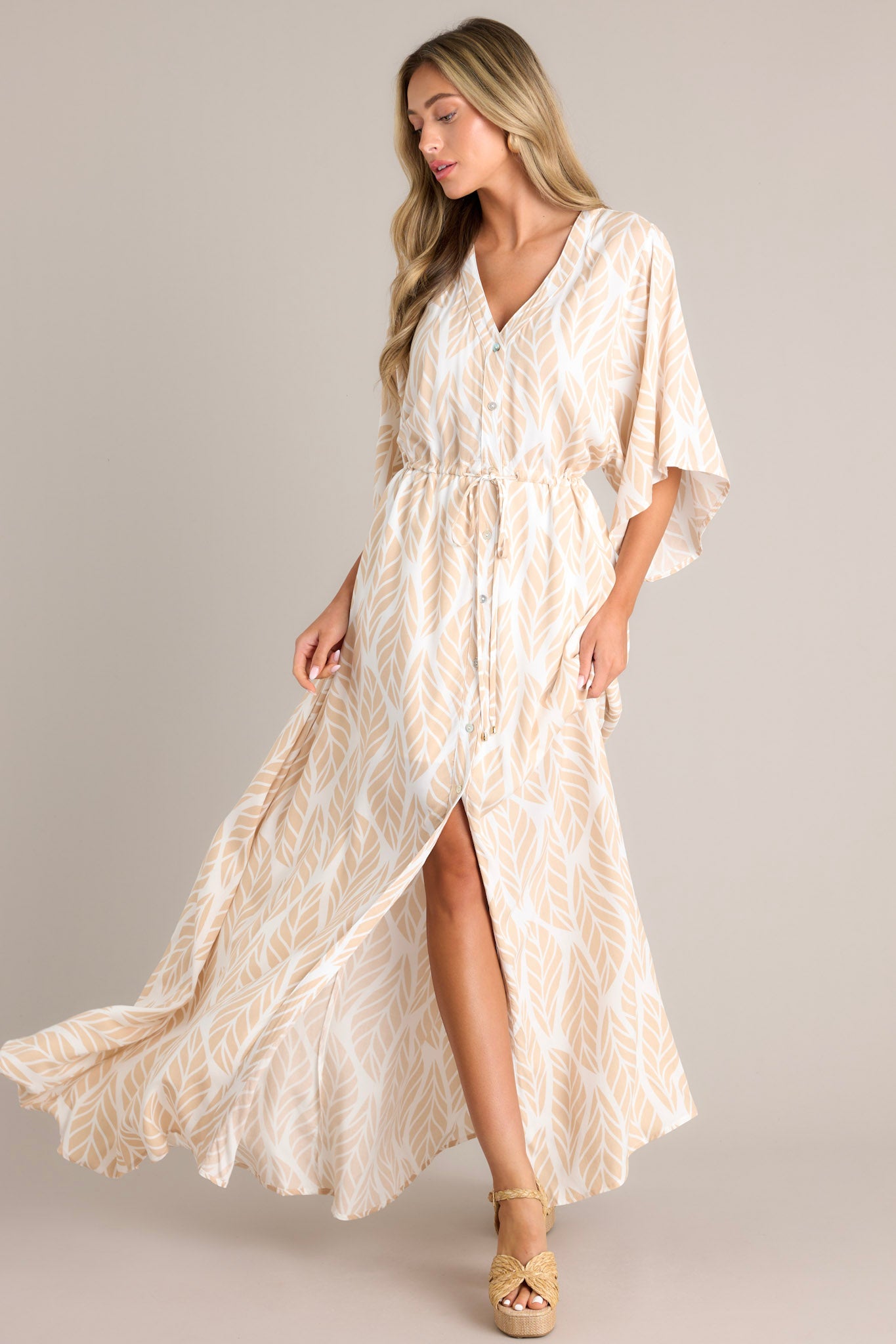 This beige maxi dress features a v-neckline, a functional button front, a self-tie drawstring waist, a flowing silhouette, and flowing half sleeves.