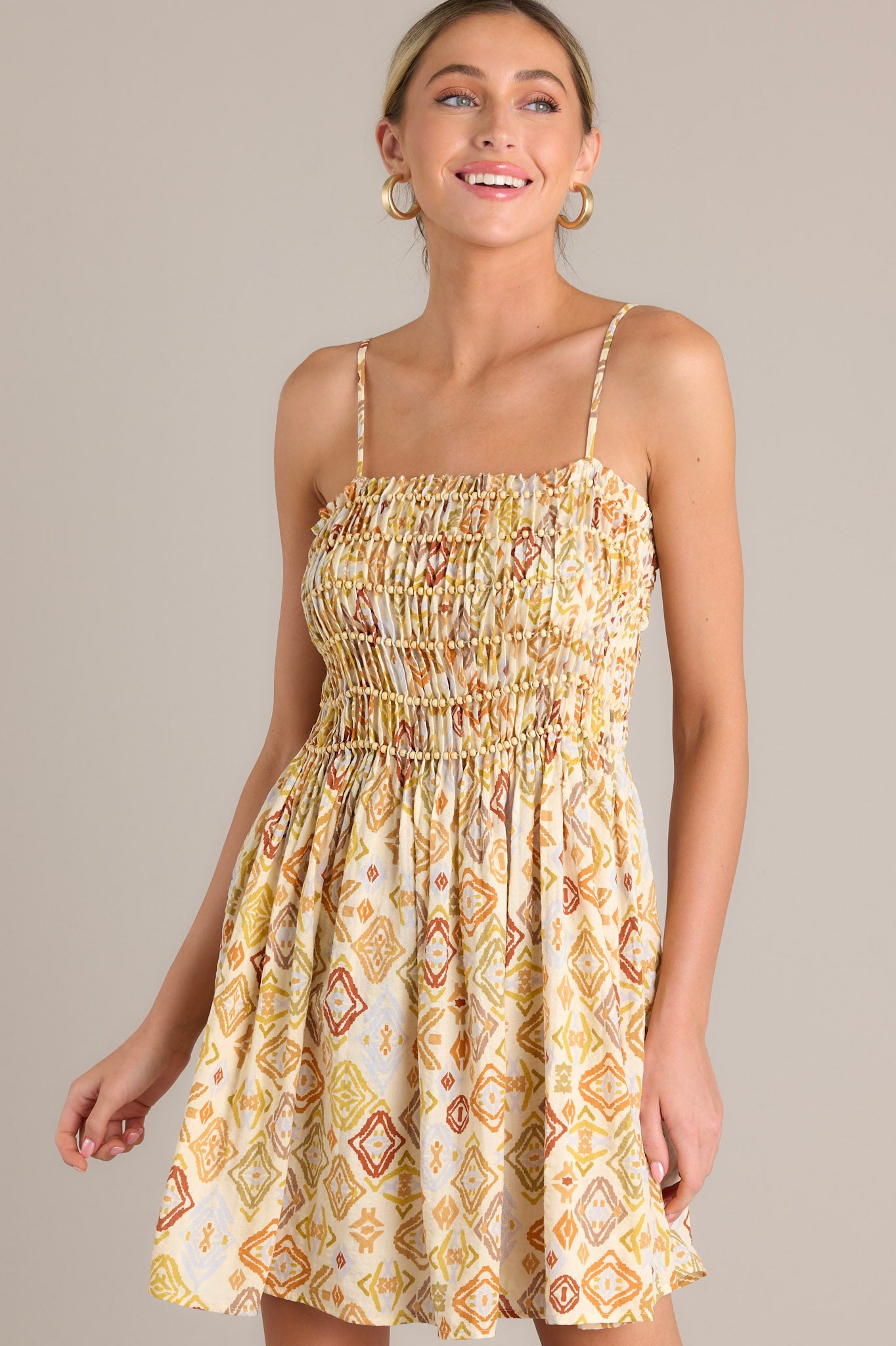 Front angled view of a beige mini dress featuring a square neckline, thin adjustable straps, fully smocked pleated bodice with beaded detailing, and a flowing skirt