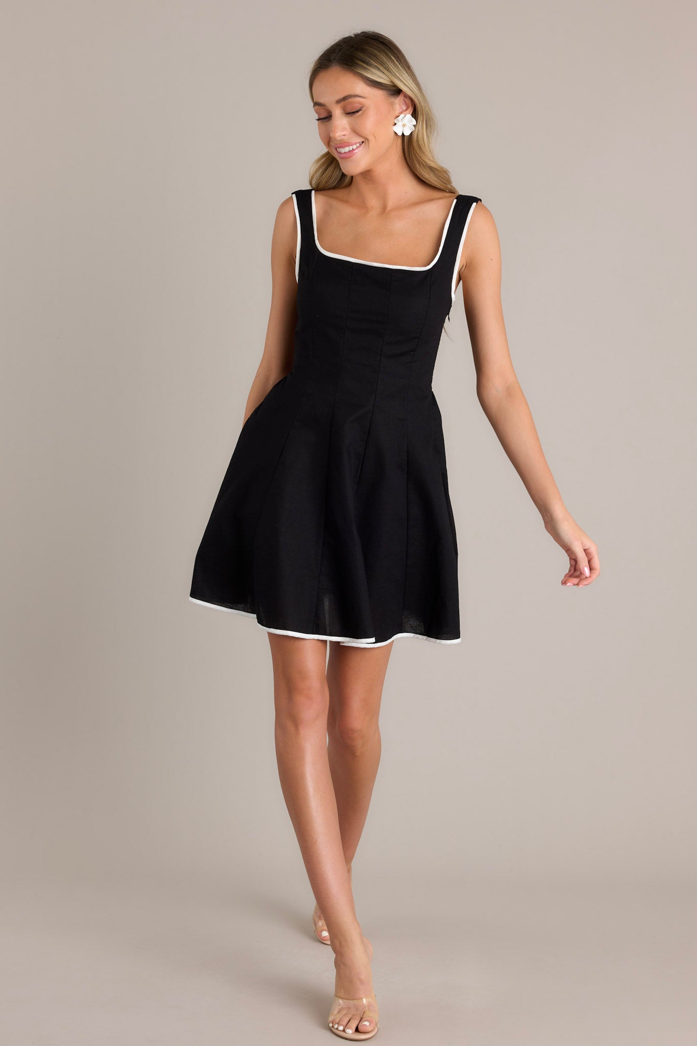 Movement shot of a black mini dress with a square neckline, thick adjustable straps, a discrete side zipper, a smocked back insert, and contrasting white trim, capturing the flow and fit of the dress as it moves.