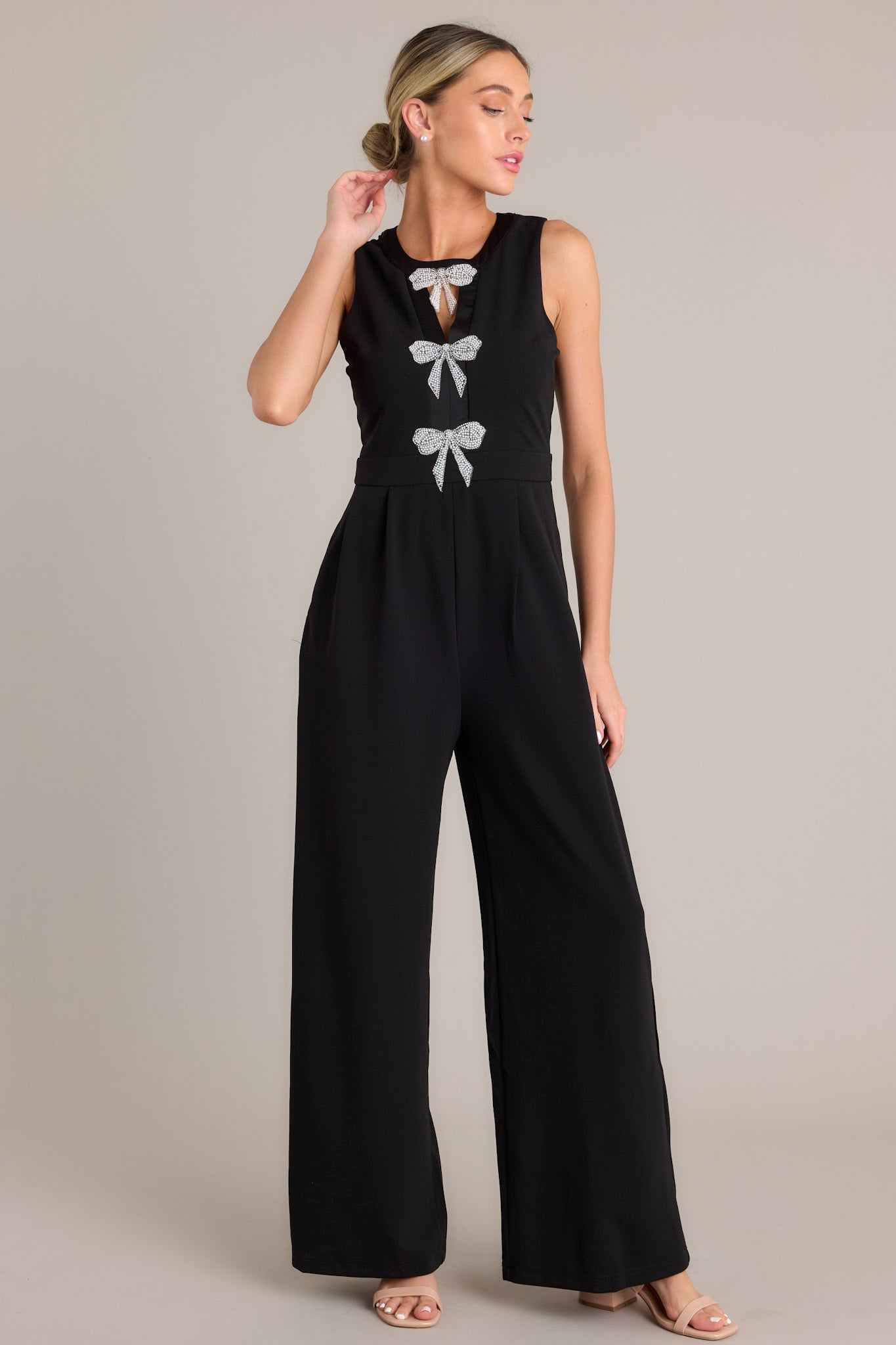 This black jumpsuit features a round neckline, a discrete back zipper, three ornate bows embellished with faux ivory pearls and rhinestones, a chest cutout, a thick waistband, subtle pleats, functional hip pockets, and a wide leg design.