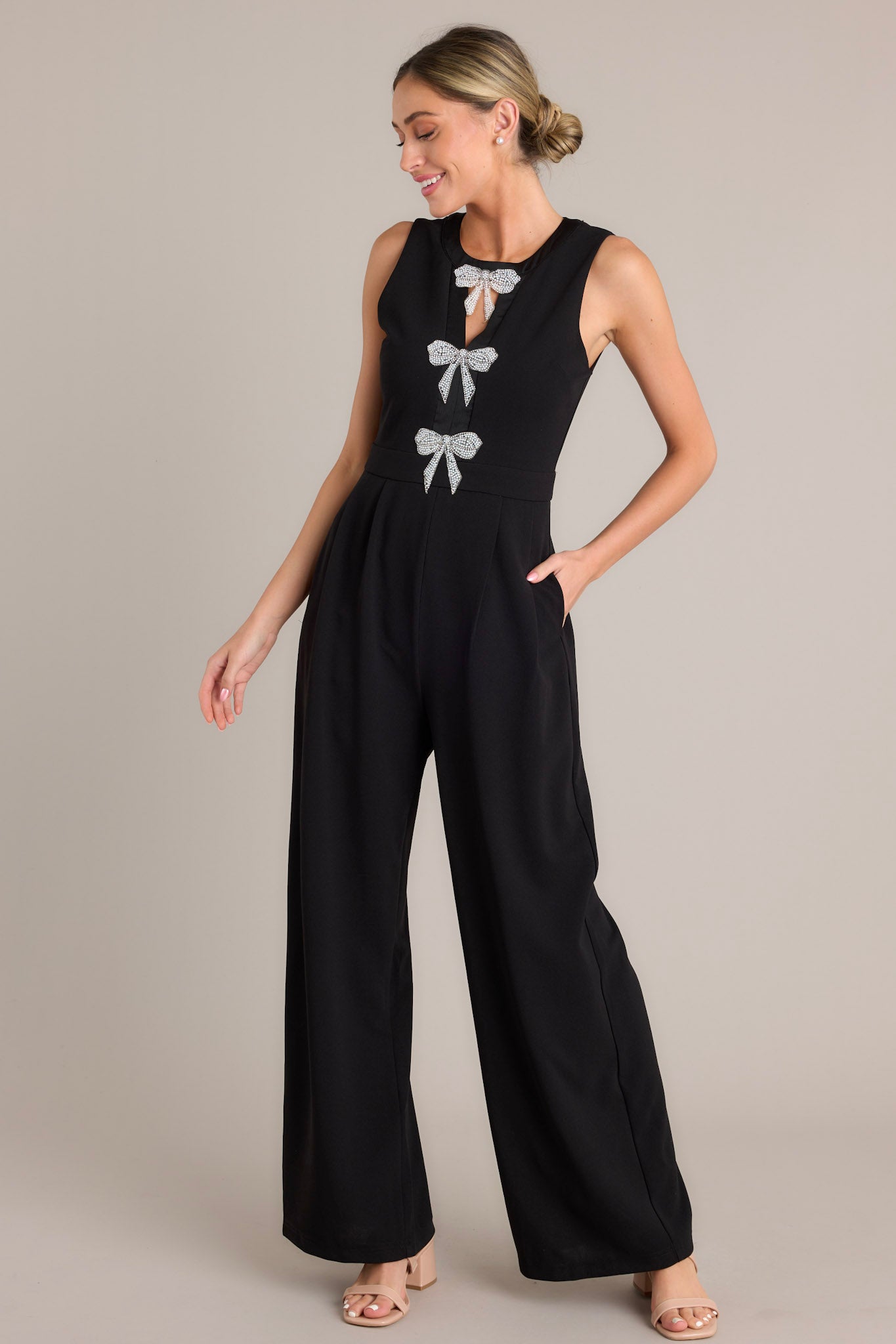 Angled full body view of this black jumpsuit that features a round neckline, a discrete back zipper, three ornate bows embellished with faux ivory pearls and rhinestones, a chest cutout, a thick waistband, subtle pleats, functional hip pockets, and a wide leg design.