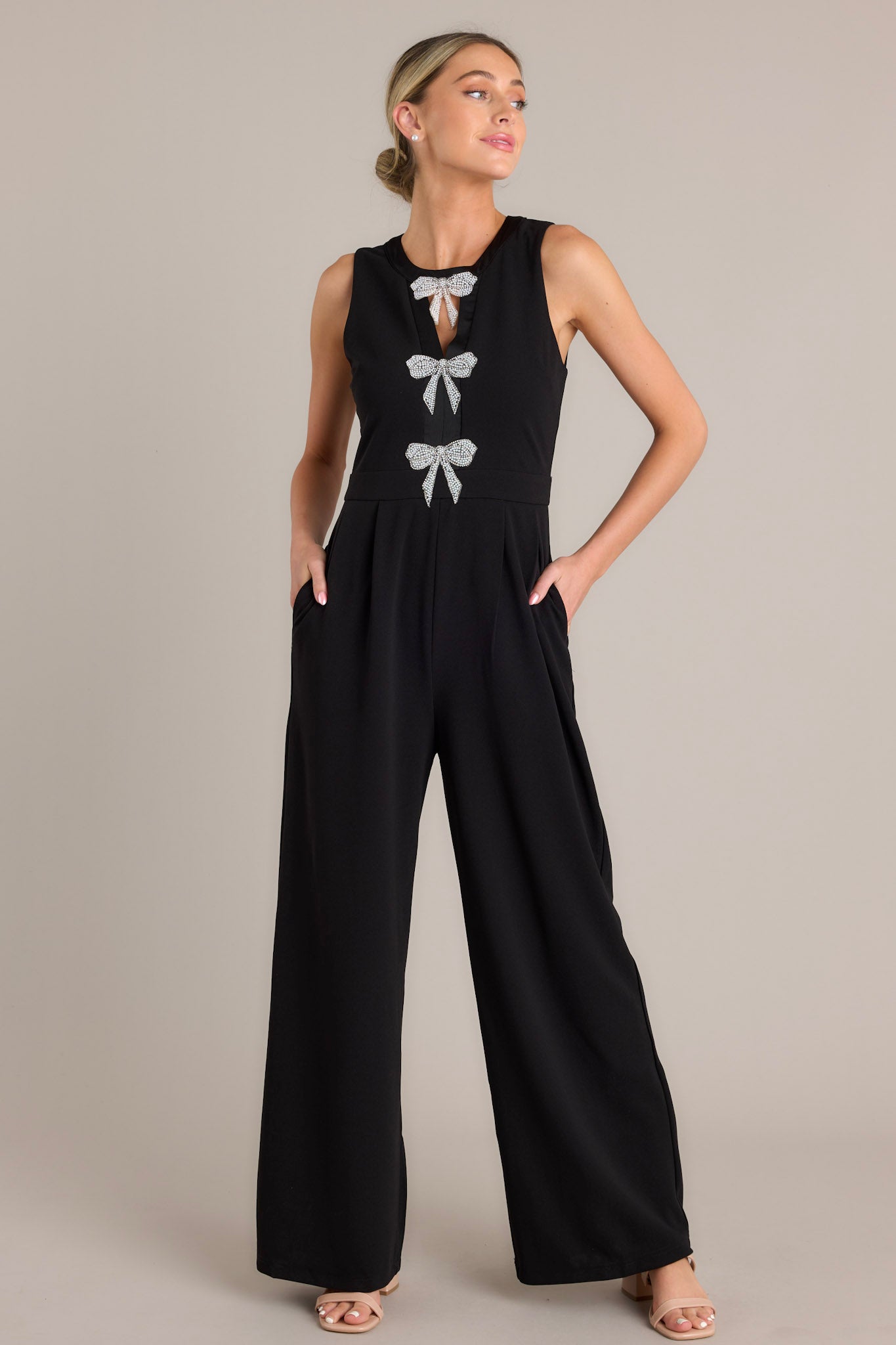 Front view of this black jumpsuit that features a round neckline, a discrete back zipper, three ornate bows embellished with faux ivory pearls and rhinestones, a chest cutout, a thick waistband, subtle pleats, functional hip pockets, and a wide leg design.