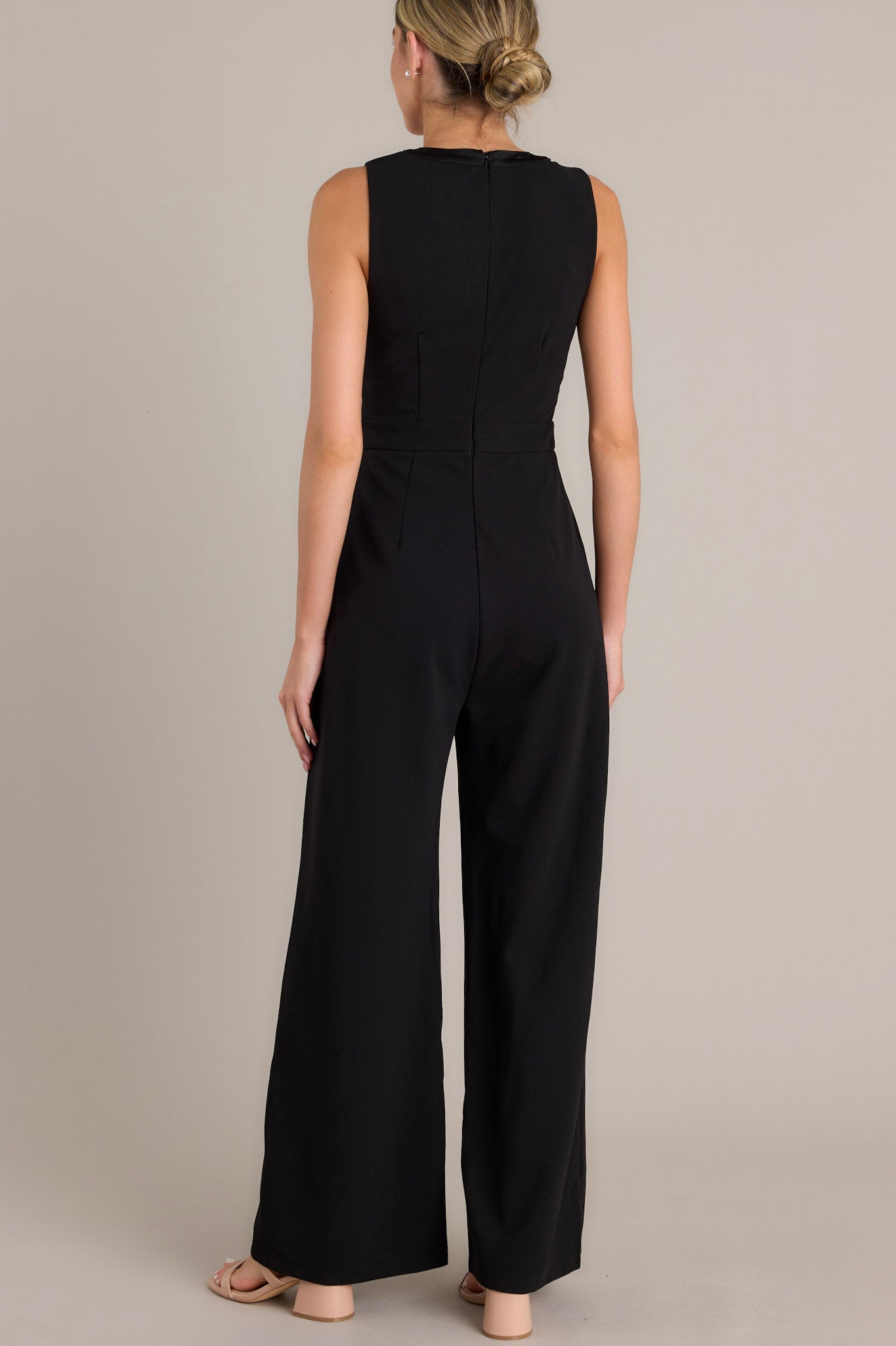 Back view of this black jumpsuit that features a round neckline, a discrete back zipper, three ornate bows embellished with faux ivory pearls and rhinestones, a chest cutout, a thick waistband, subtle pleats, functional hip pockets, and a wide leg design.