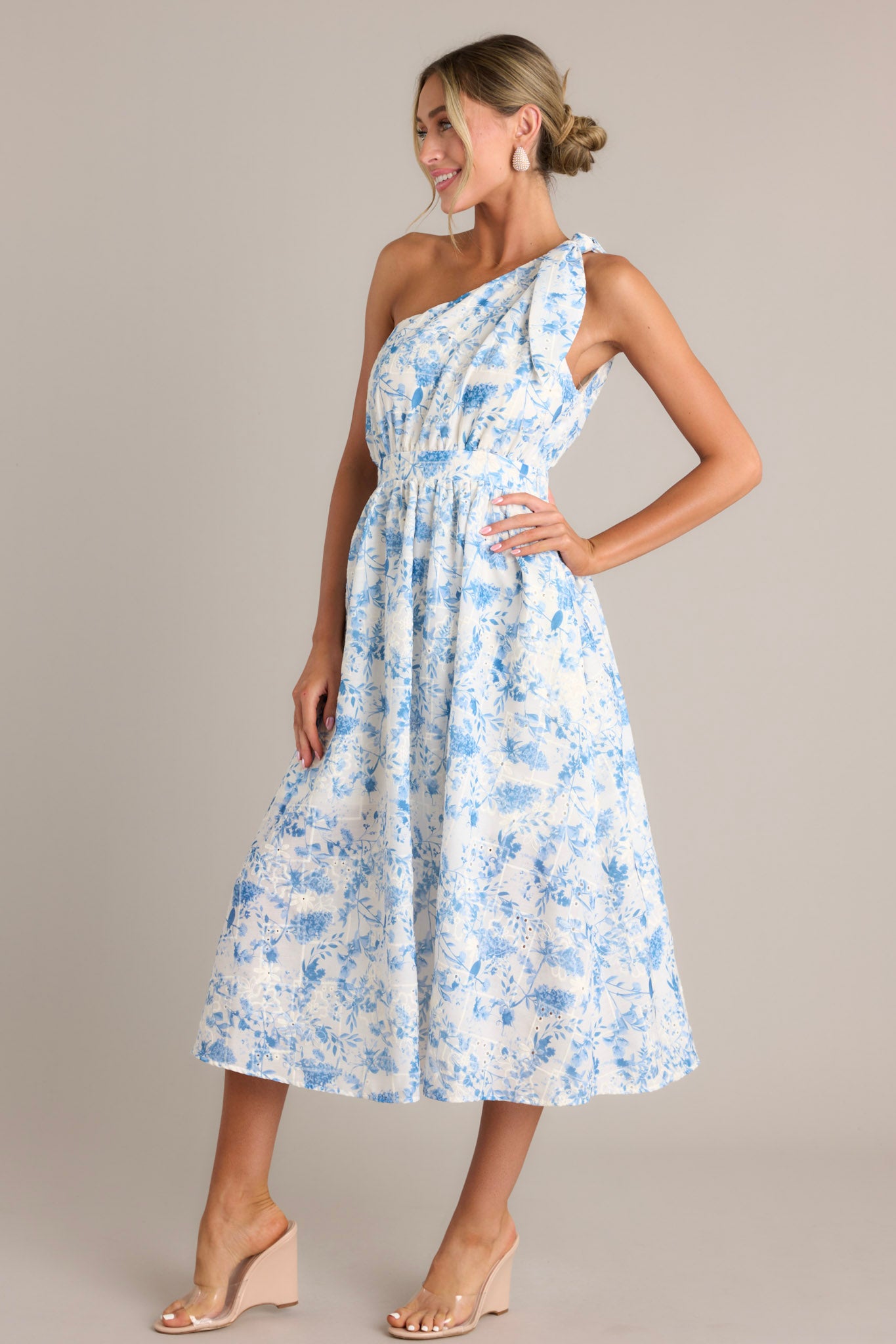 This blue floral dress features an asymmetrical neckline, self-tie shoulder feature, a thick waistband with an elastic insert, a discrete side zipper, functional hip pockets, and embroidered details throughout.