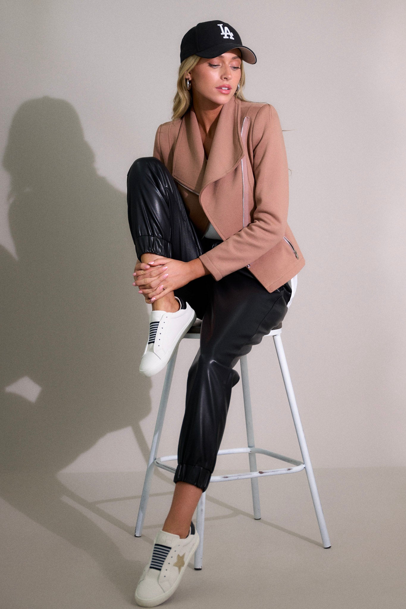 Seated Front angled view of a mocha tan moto jacket featuring shoulder pads, a cross zipper closure, functional waist pockets with zipper closure, lapels, and silver hardware