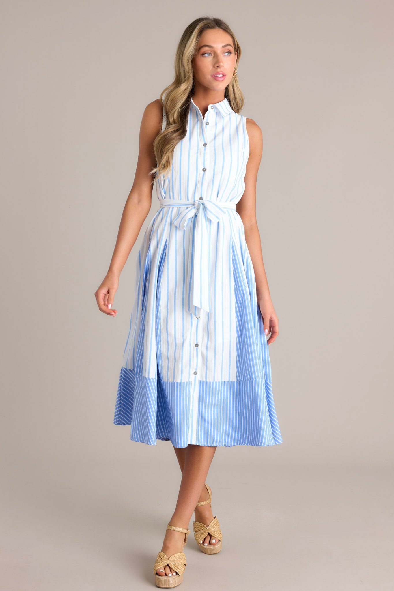 Front angled view of a light blue midi dress featuring a collared neckline, a functional button front, a self-tie waist belt, a unique stripe pattern, and a flowing silhouette