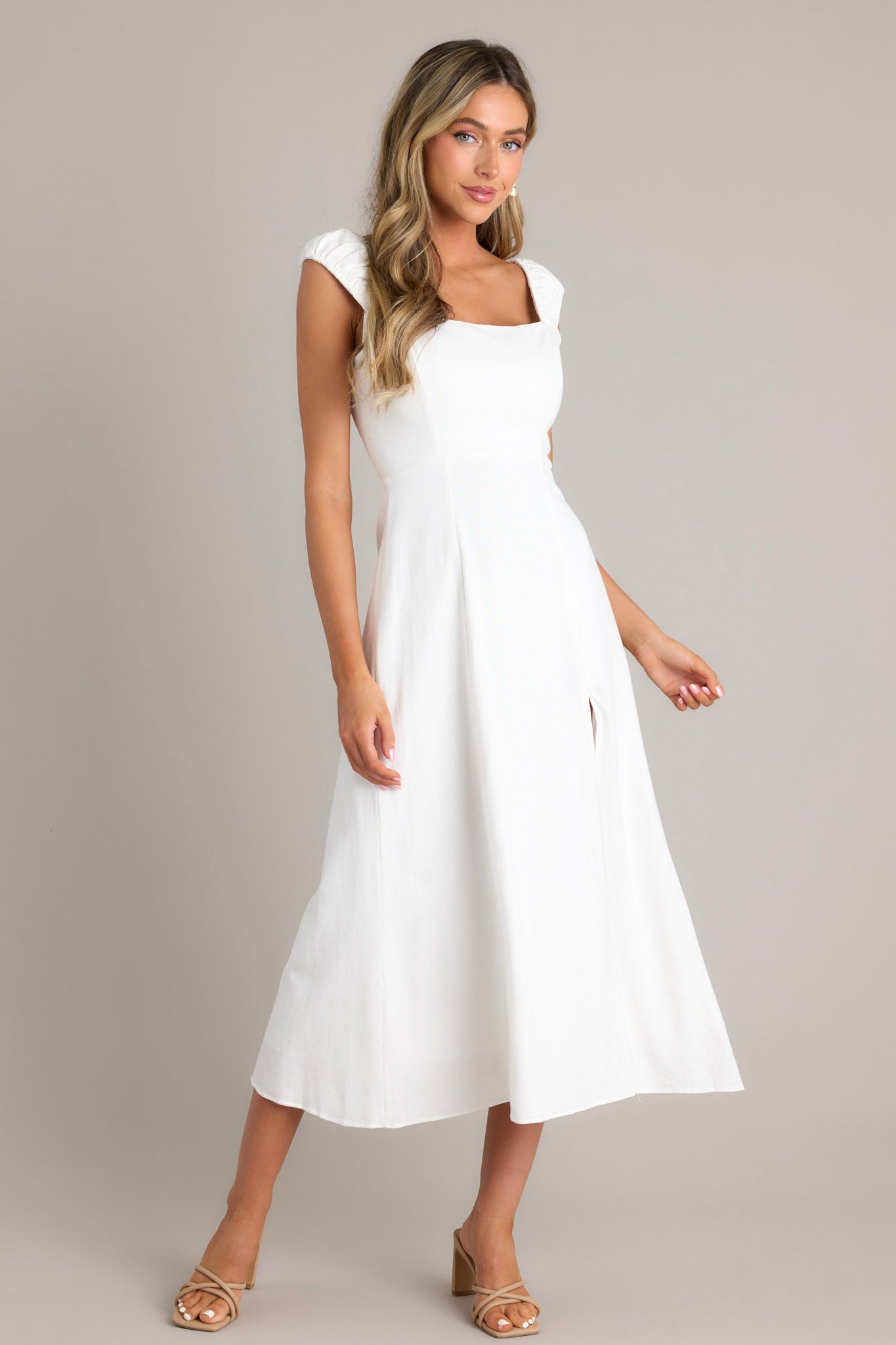 Front angled view of a white midi dress featuring a square neckline, smocked back section, hidden zipper, smocked sleeves, leg slit, and flowy skirt