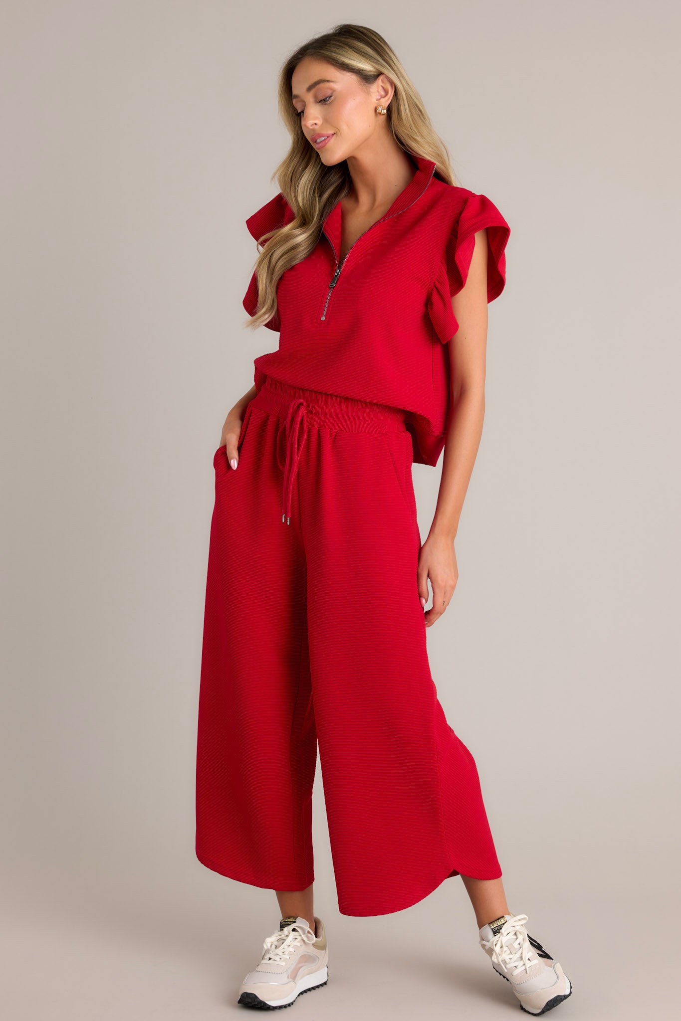 Front angled view of red pants displaying the high waisted design, elastic waistband, self-tie drawstring, functional hip pockets, ribbed texture, and wide leg with a scooped hemline, worn by a model.