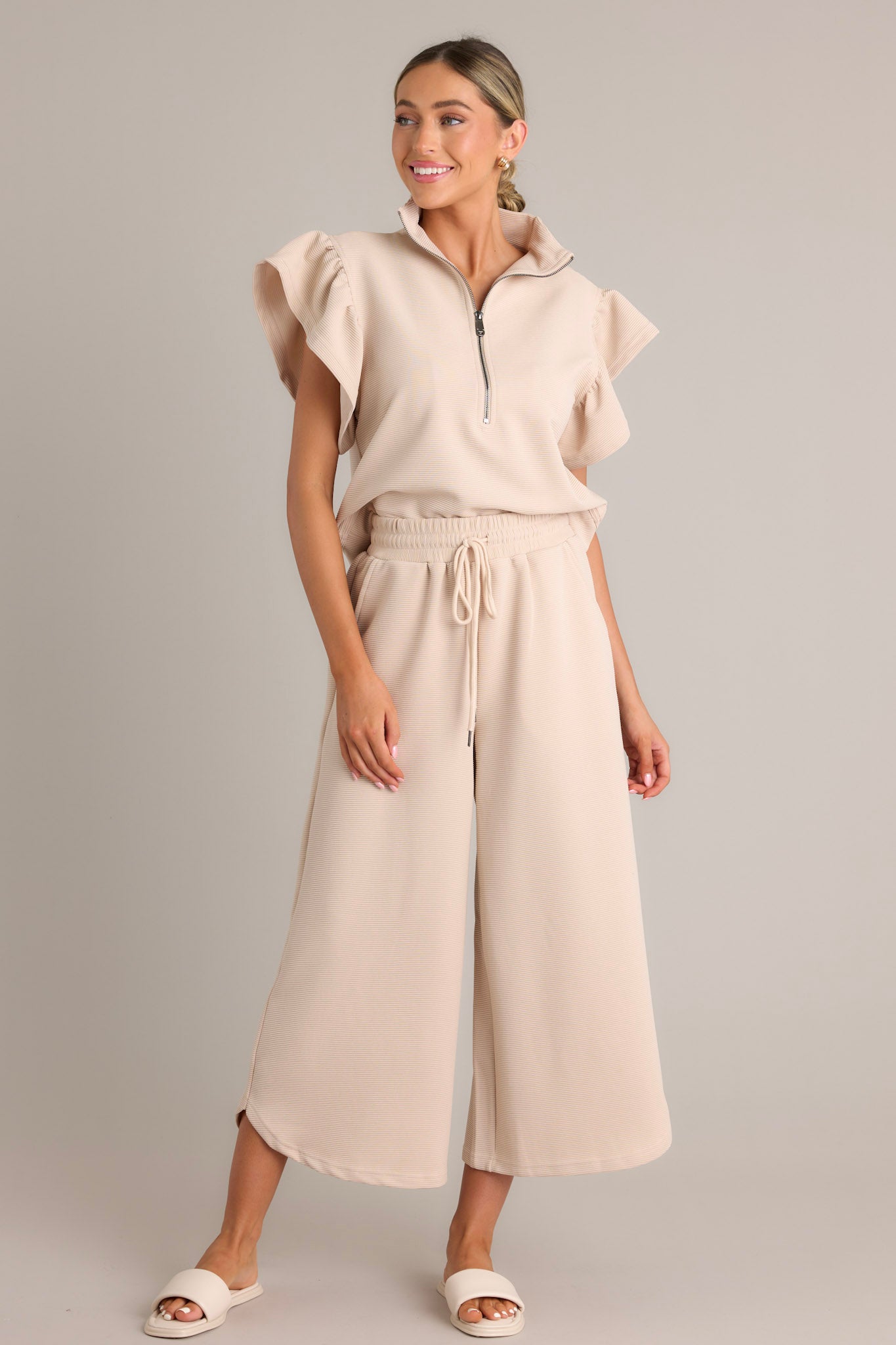 Full length view of tan pants featuring a high waisted design, elastic waistband, self-tie drawstring, functional hip pockets, ribbed texture, wide leg, and a scooped hemline.