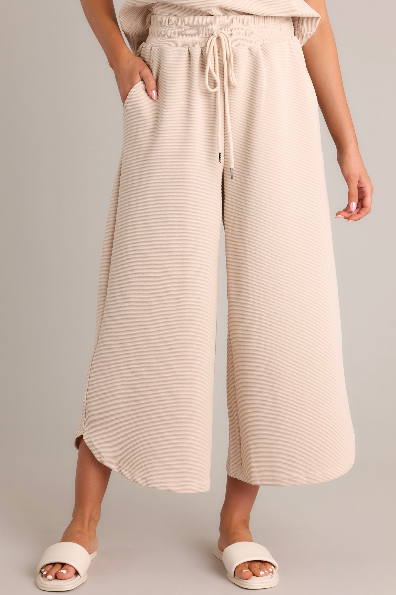 Front view of tan pants featuring a high waisted design, elastic waistband, self-tie drawstring, functional hip pockets, ribbed texture, wide leg, and a scooped hemline.