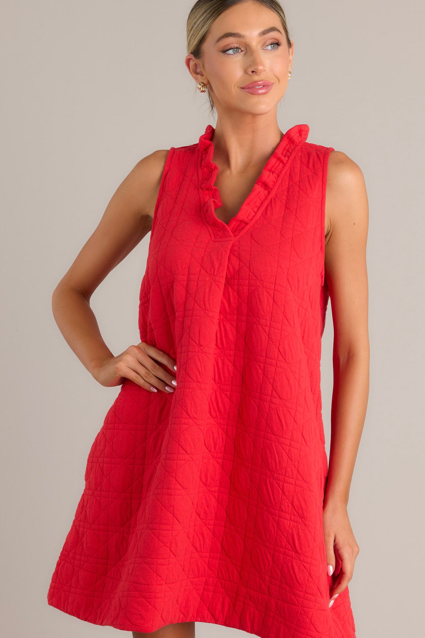 Front angled view of a red mini dress featuring a ruffled v-neckline, unique quilted material, functional hip pockets, and a sleeveless design