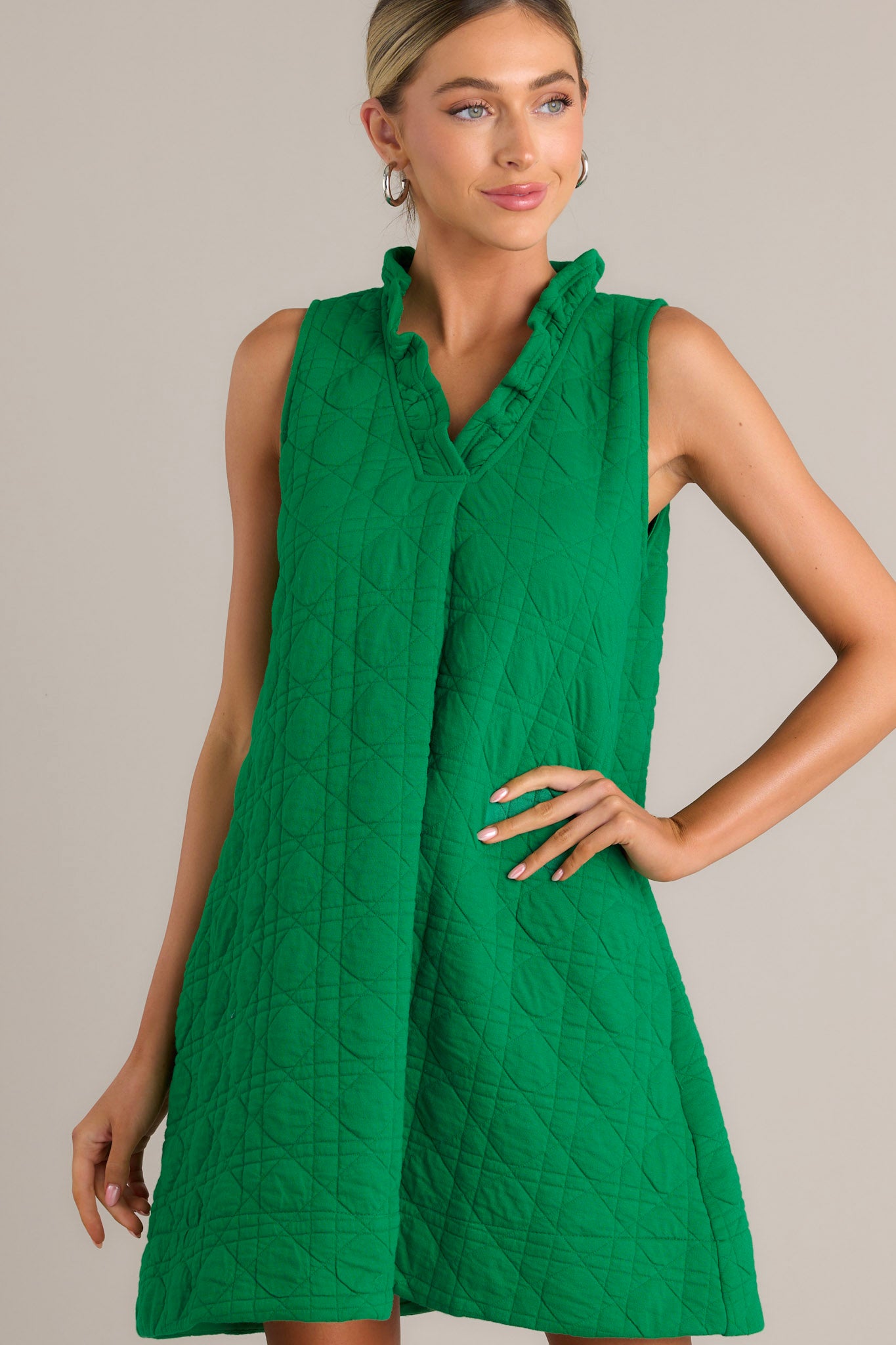 Front angled view of a green mini dress featuring a ruffled v-neckline, a unique quilted material, functional hip pockets, and a sleeveless design