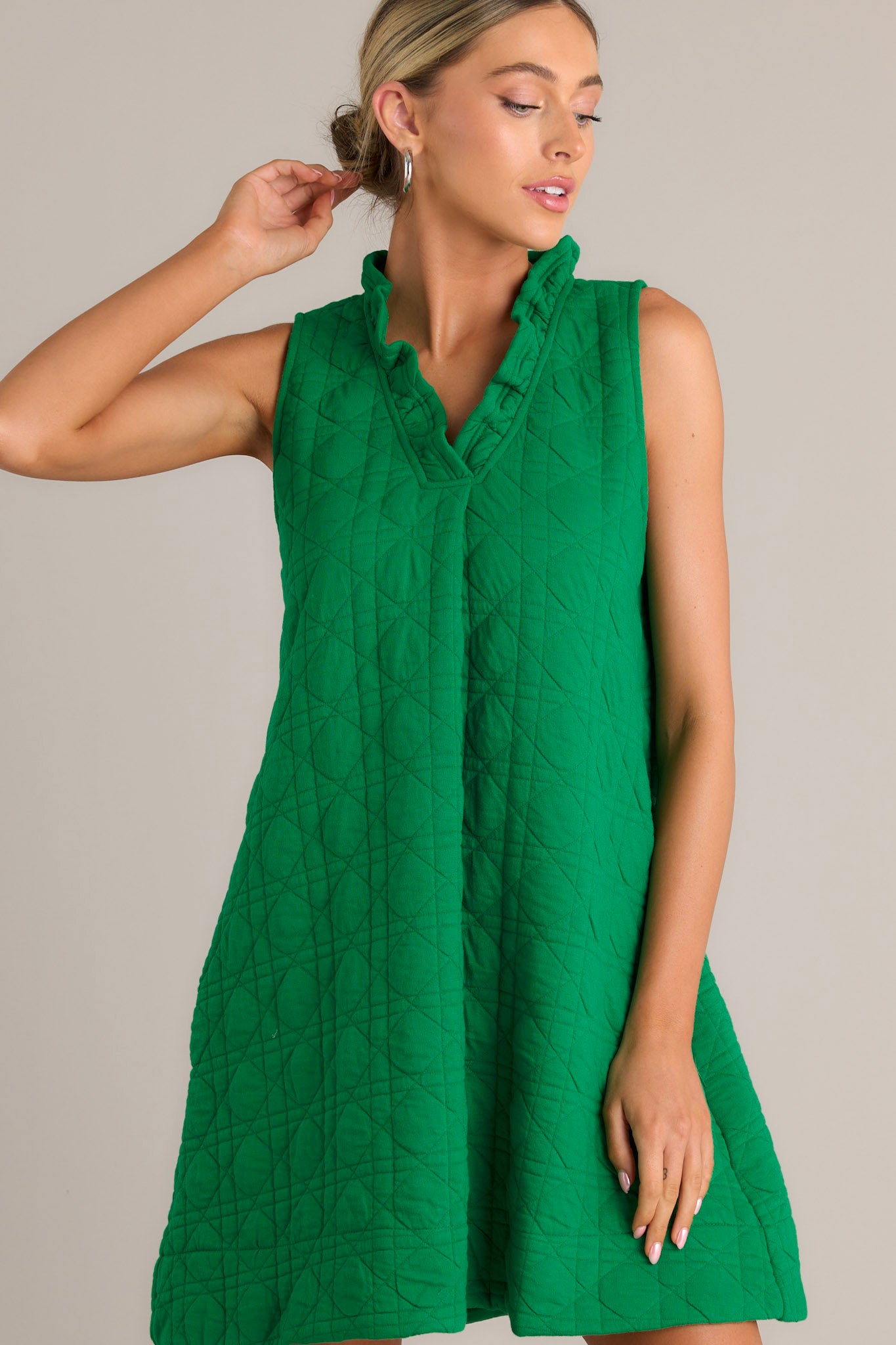 Front view of a green mini dress featuring a ruffled v-neckline, a unique quilted material, functional hip pockets, and a sleeveless design.