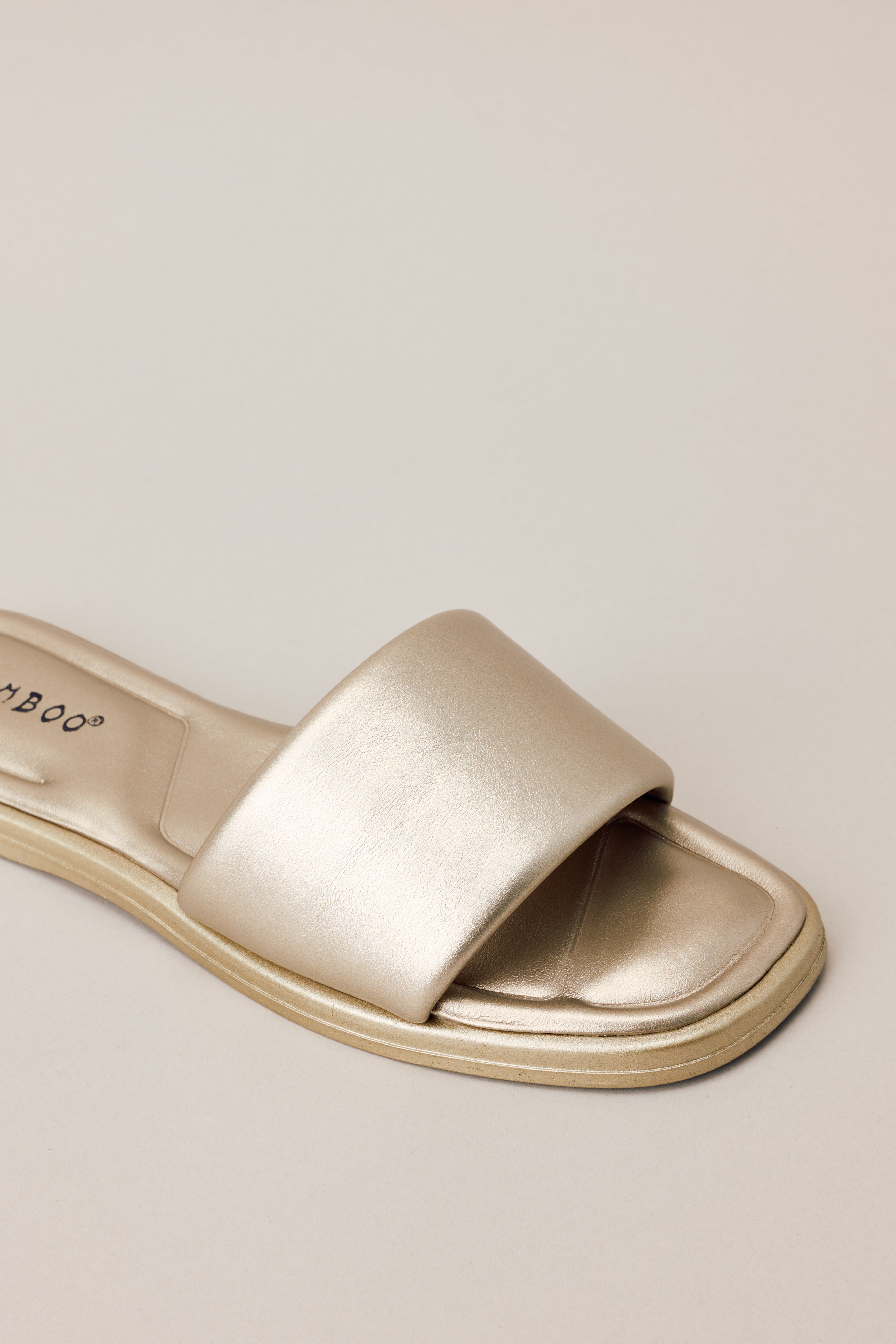 Angled overhead view of these gold sandals that feature a square toe, a slip on design, a thick strap over the top of the foot, and cushioned soles.