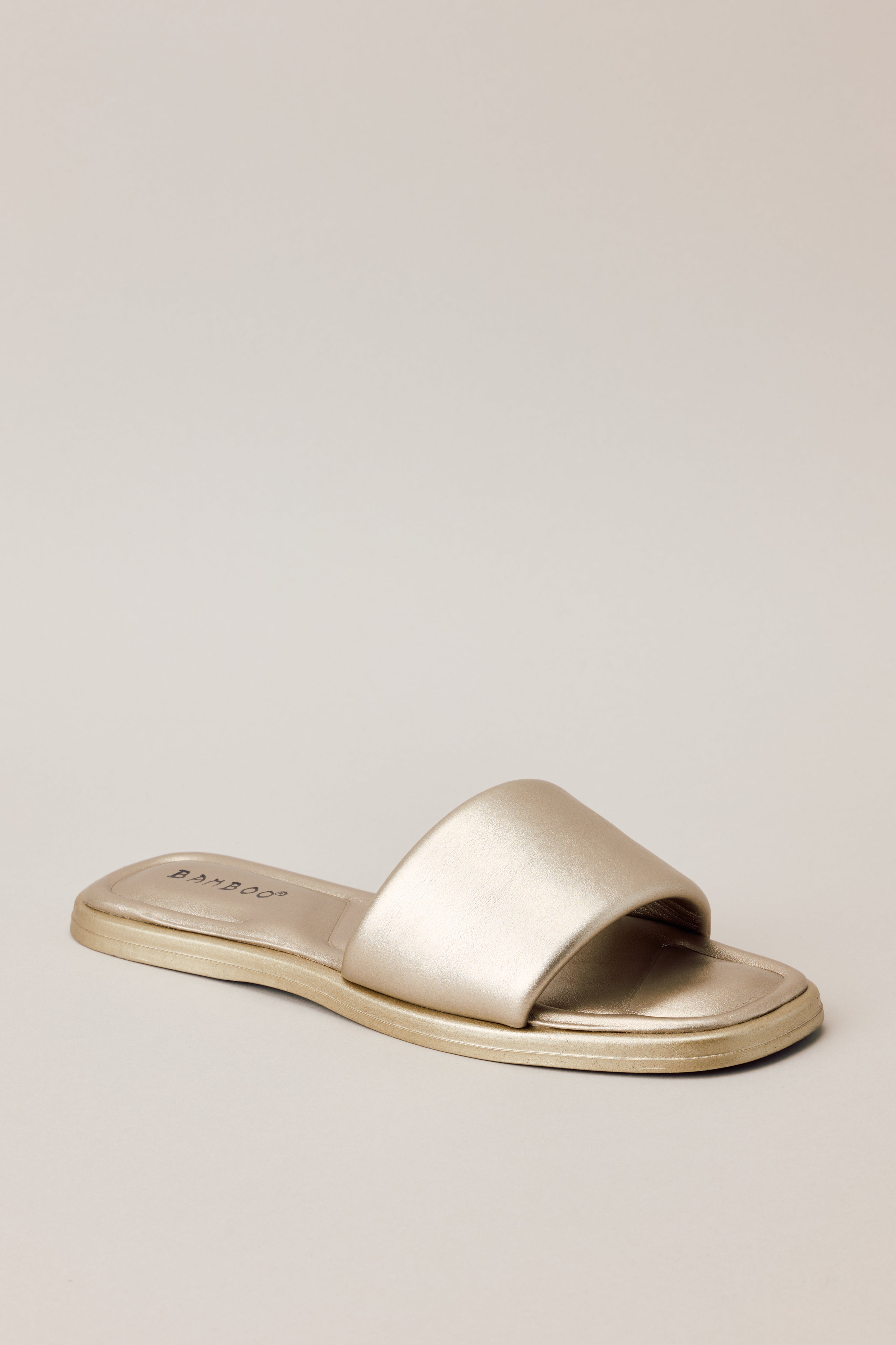 Front angled view of these gold sandals that feature a square toe, a slip on design, a thick strap over the top of the foot, and cushioned soles.