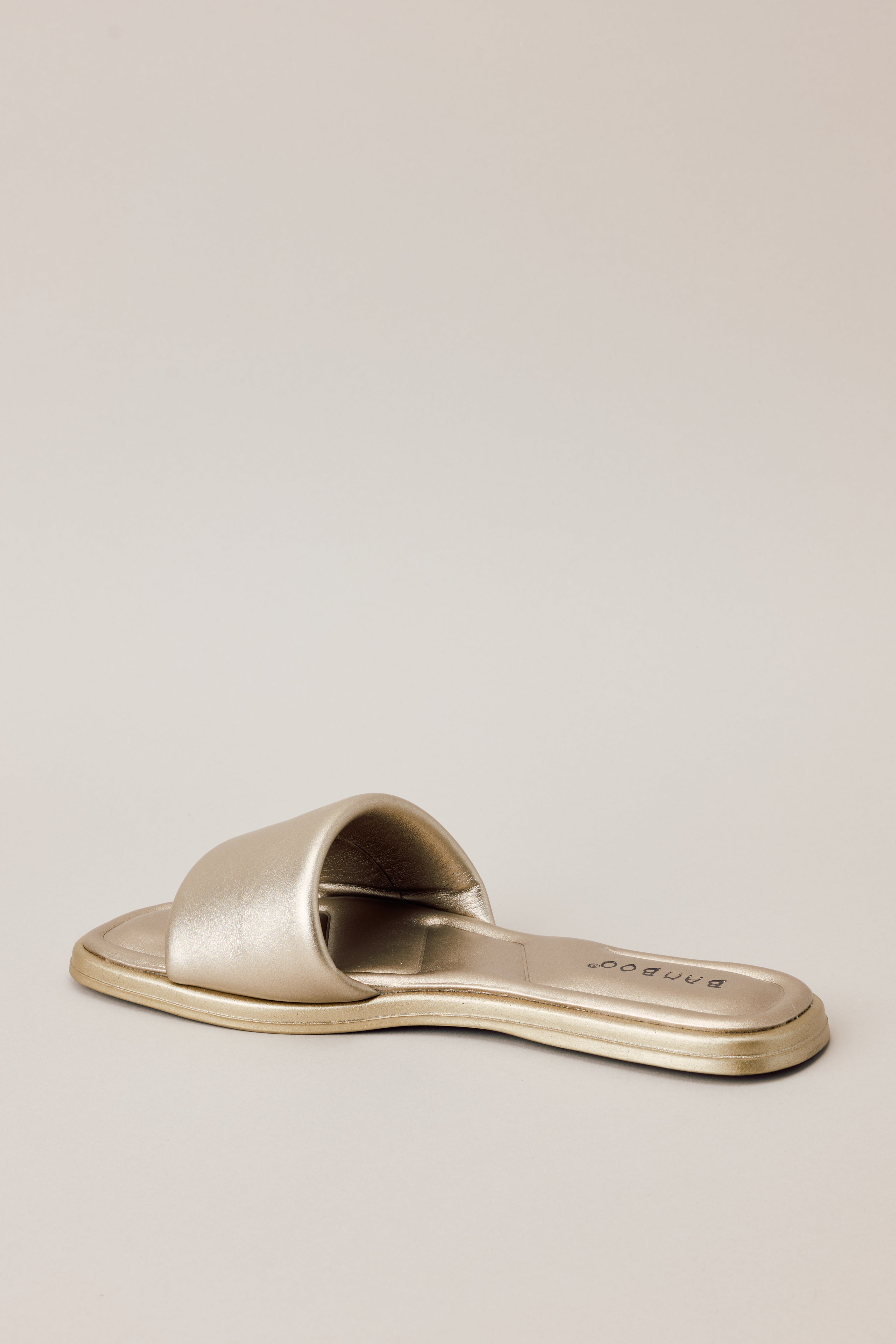 Angled back view of these gold sandals feature a square toe, a slip on design, a thick strap over the top of the foot, and cushioned soles.