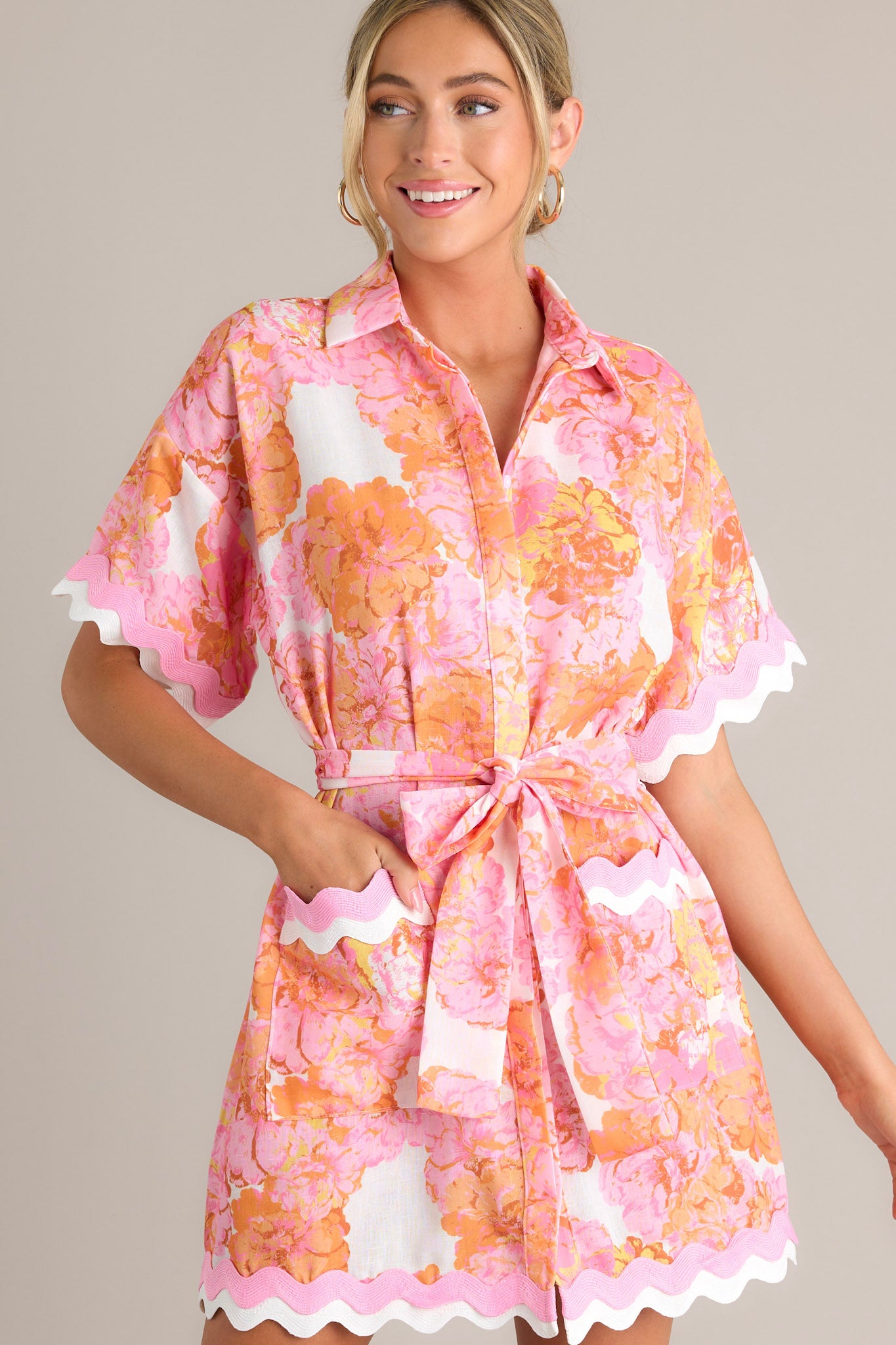 Front angled view of a light pink floral mini dress featuring a collared neckline, a functional button front, a self-tie waist belt, functional pockets, ricrac trim throughout, and short sleeves