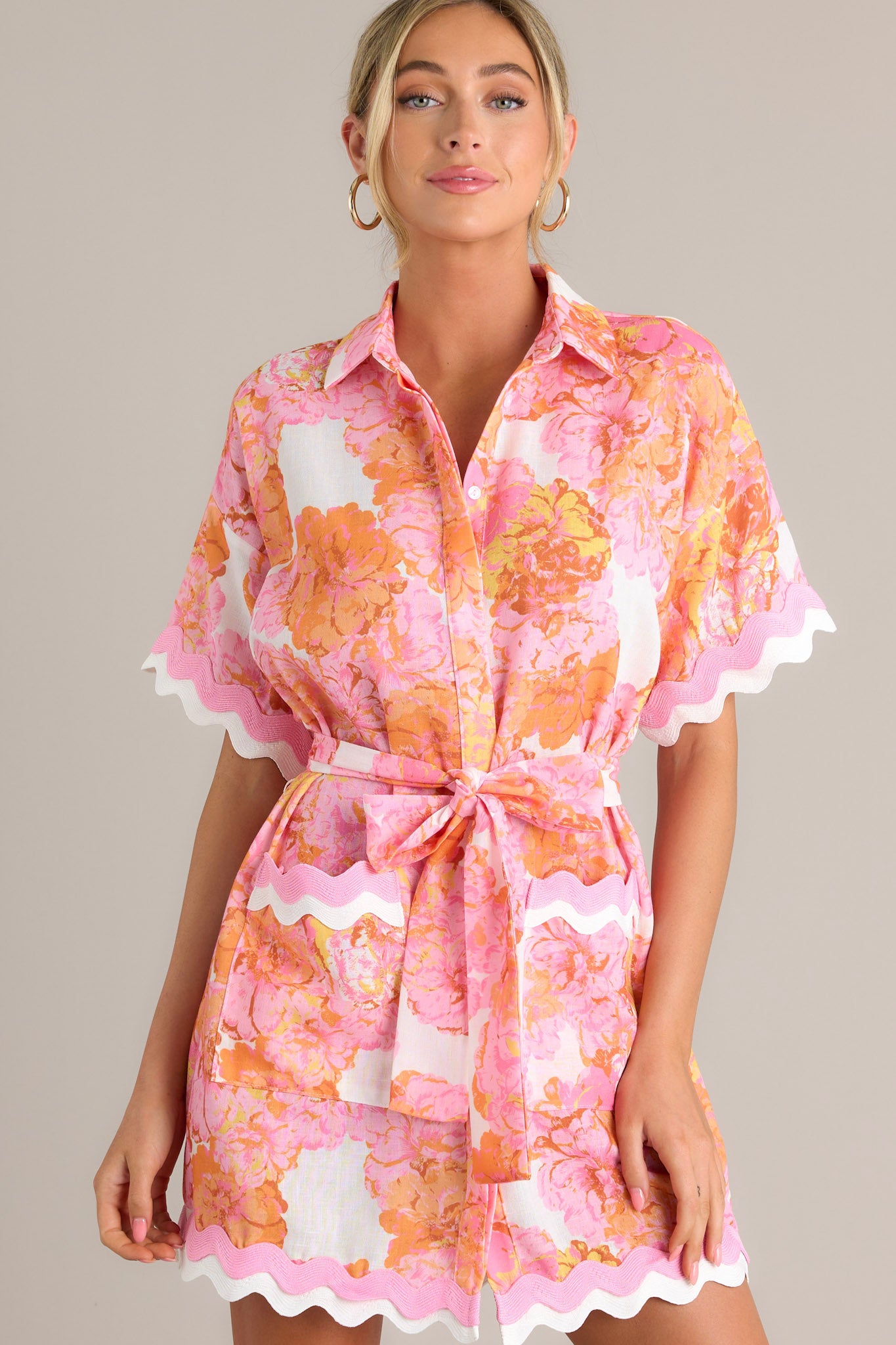 Front view of a light pink floral mini dress featuring a collared neckline, a functional button front, a self-tie waist belt, functional pockets, ricrac trim throughout, and short sleeves.