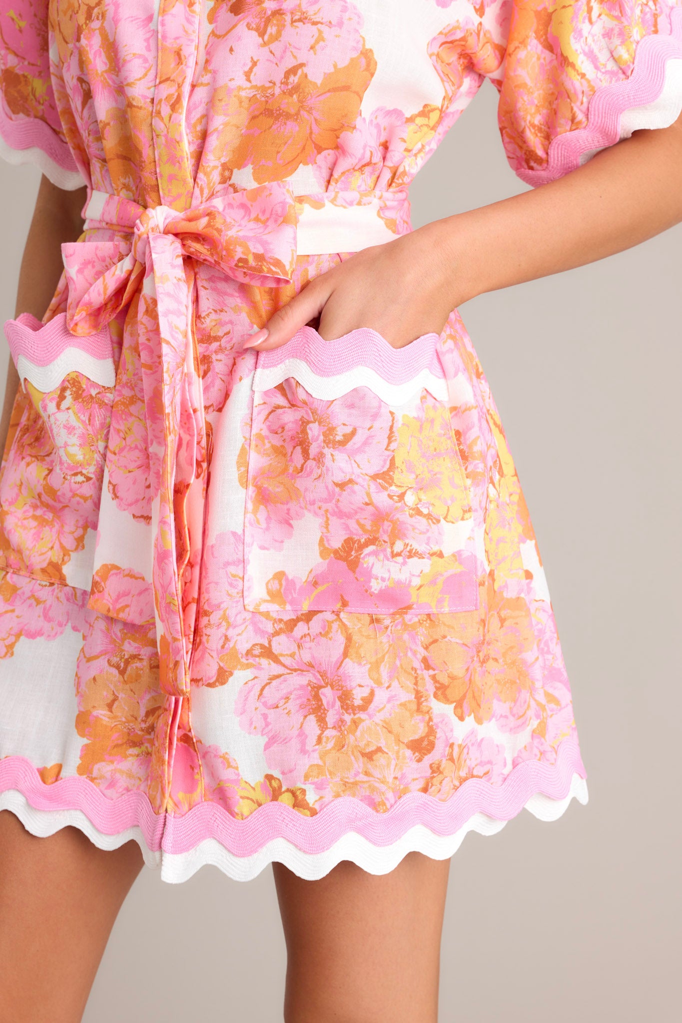 Side view of a light pink floral mini dress showcasing the collared neckline, functional button front, self-tie waist belt, functional pockets, ricrac trim throughout, and short sleeves.