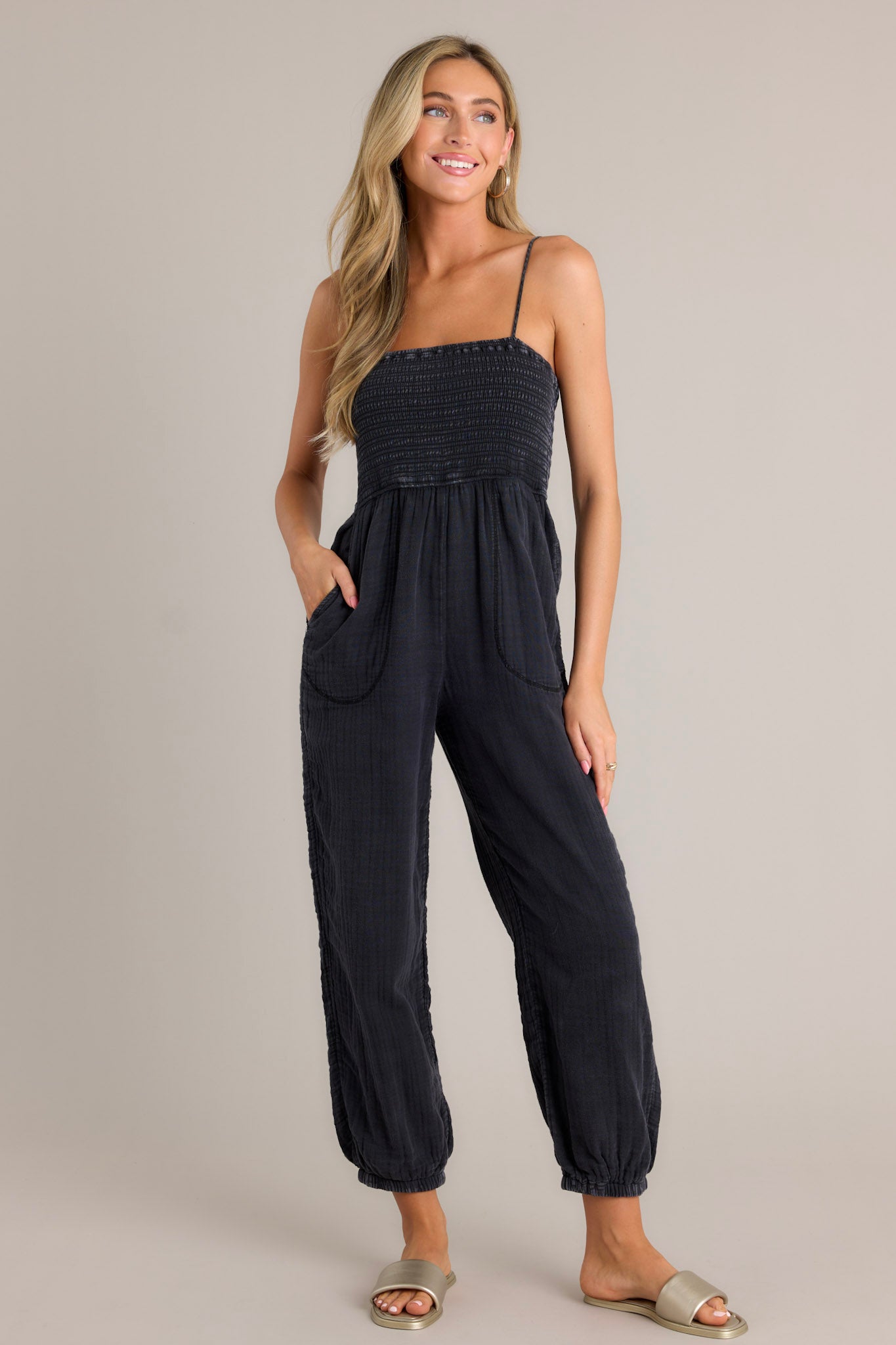 Full length view of a black jumpsuit with a square neckline, thin adjustable straps, a fully smocked bodice, functional hip pockets, a wide leg design, and elastic cuffed ankles