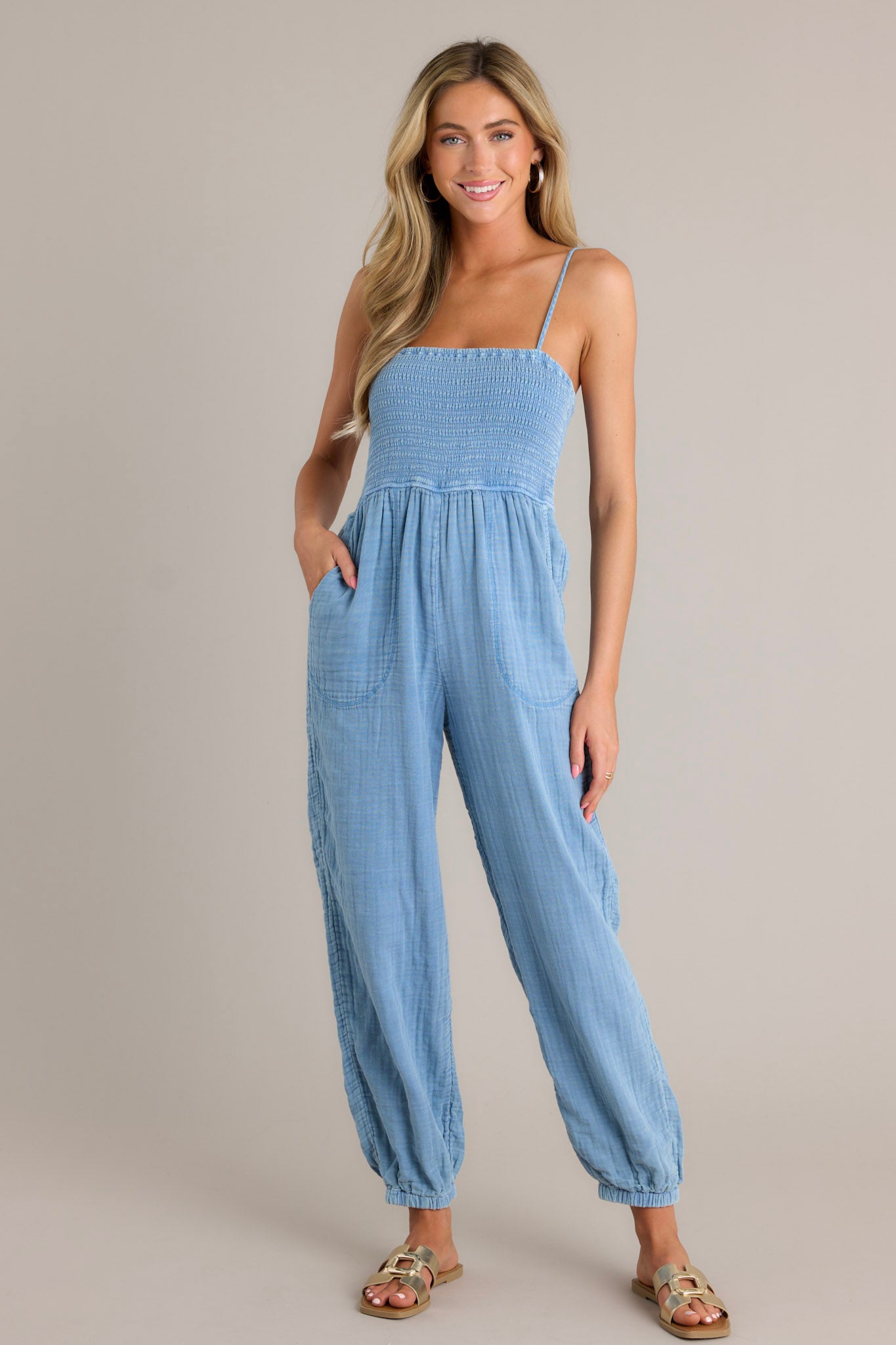 Full length view of an indigo jumpsuit with a square neckline, thin adjustable straps, a fully smocked bodice, functional hip pockets, a wide leg design, and elastic cuffed ankles.