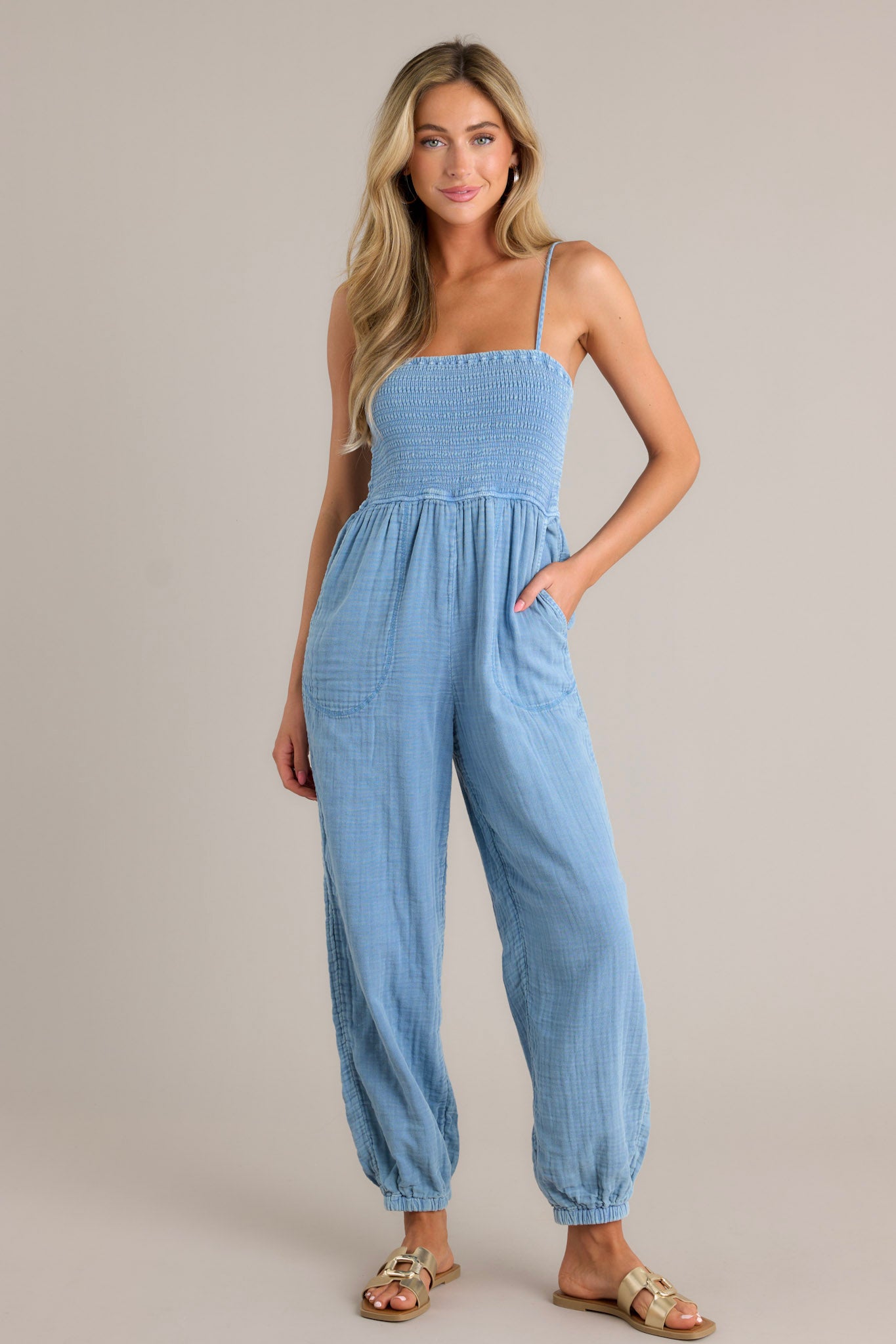 Front view of an indigo jumpsuit featuring a square neckline, thin adjustable straps, a fully smocked bodice, functional hip pockets, a wide leg design, and elastic cuffed ankles.