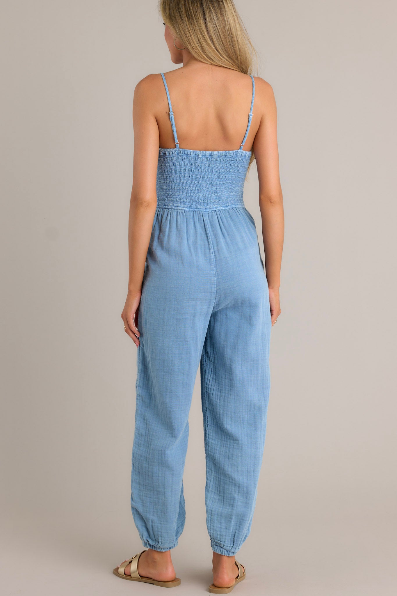 Back view of an indigo jumpsuit highlighting the thin adjustable straps, fully smocked bodice, and elastic cuffed ankles.