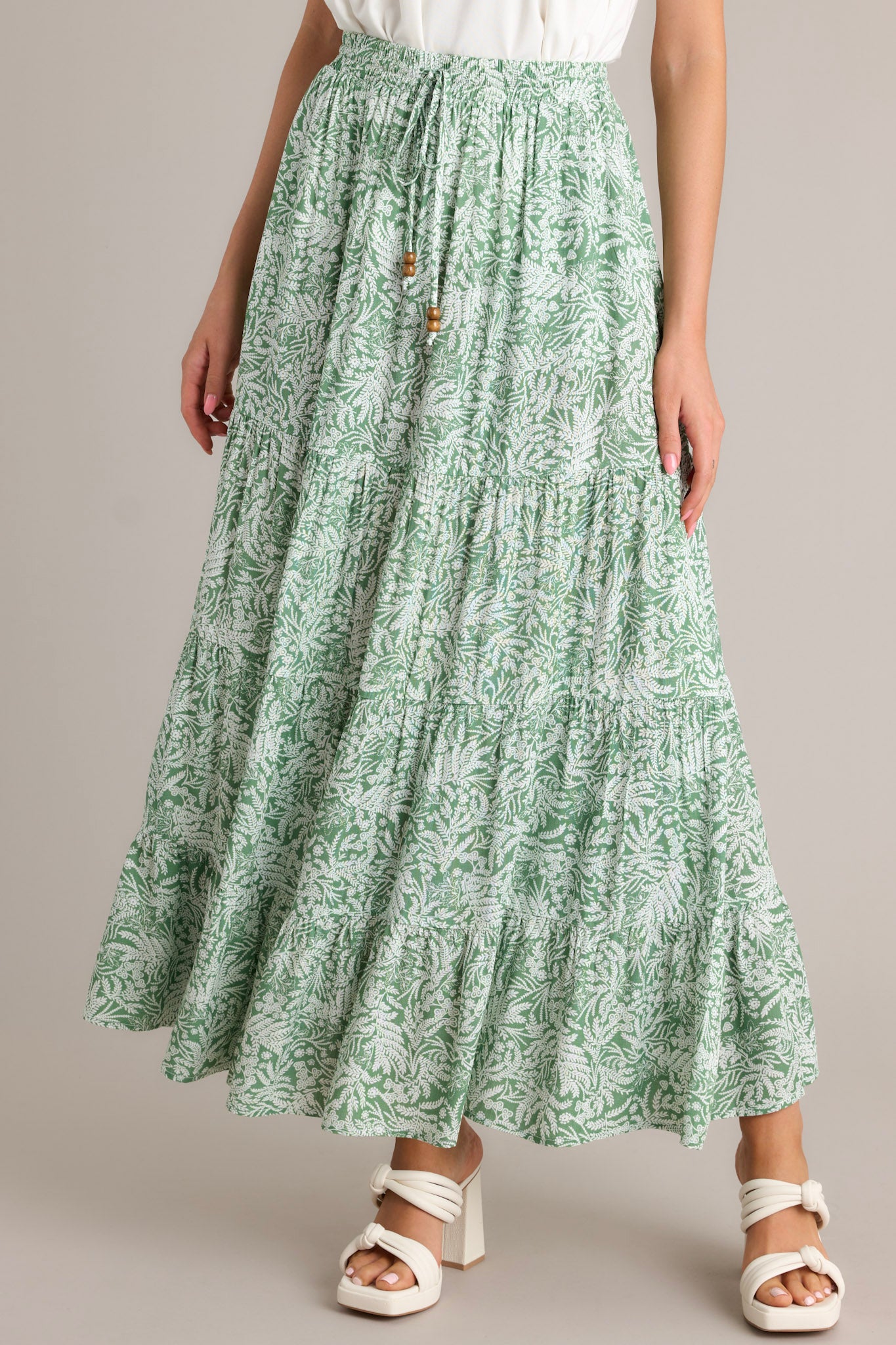 Front view of a green maxi skirt featuring a high waisted design, an elastic waistband, a self-tie drawstring, multi tiers, and a flowing silhouette.