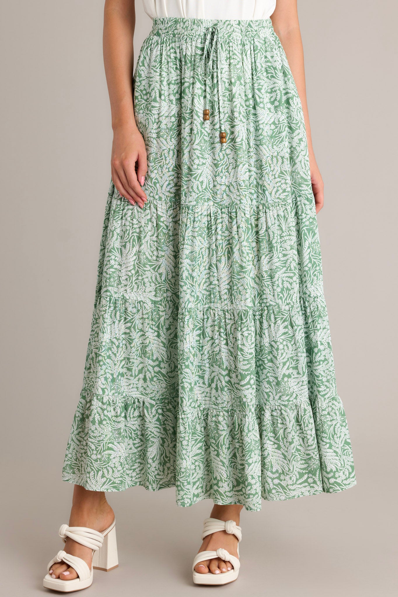 Front angled view of a green maxi skirt featuring a high waisted design, an elastic waistband, a self-tie drawstring, multi tiers, and a flowing silhouette