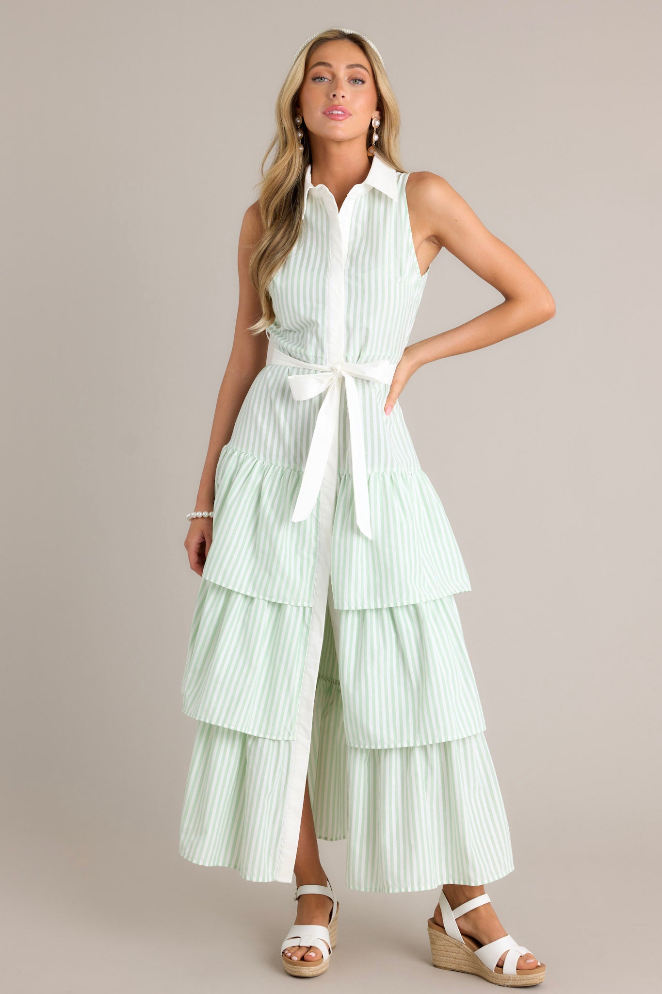 Front angled view of a mint green stripe maxi dress featuring a collared neckline, a button front, an elastic waist insert, a self-tie waist belt, and multiple tiers