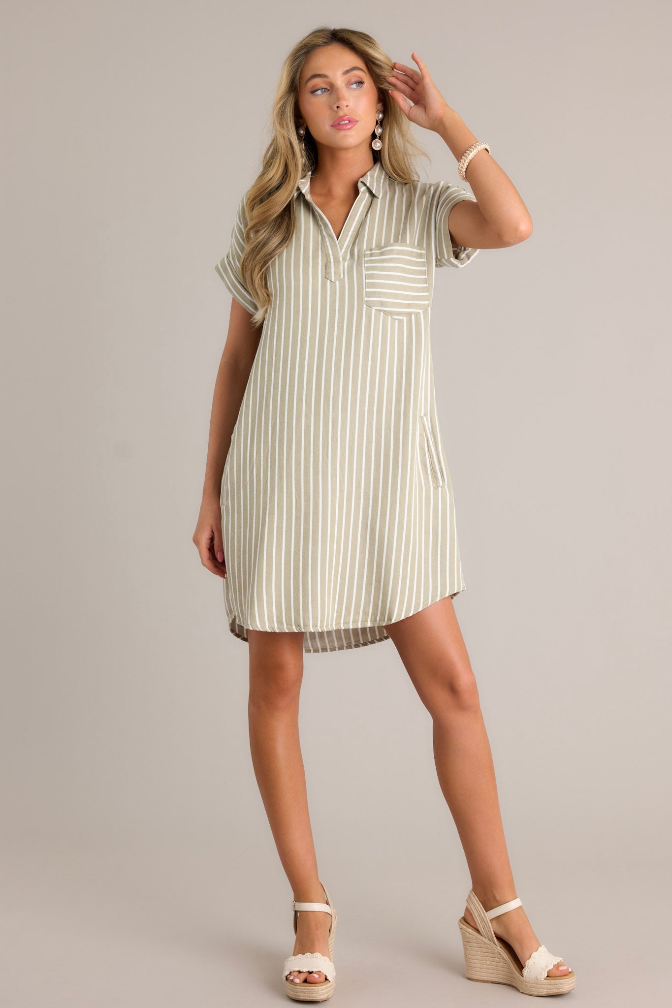 Front angled view of an olive green dress featuring a collared v-neckline, a functional breast pocket, a relaxed fit, functional hip pockets, and a scoop hemline