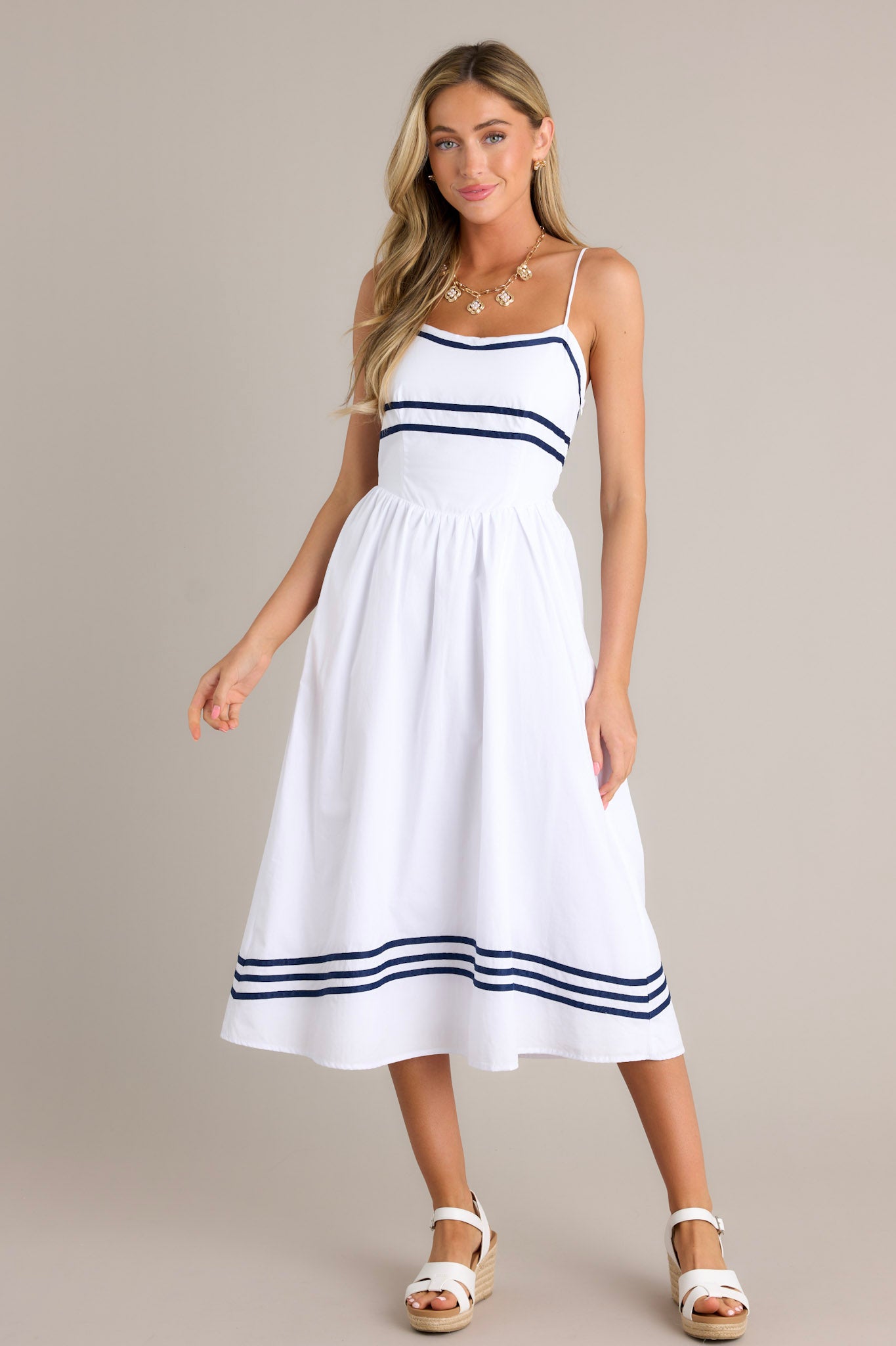 Full length view of a white midi dress with a rounded neckline, thin adjustable straps, a smocked back insert, a discrete side zipper, a dropped waist, navy stripe detailing, and a flowing silhouette