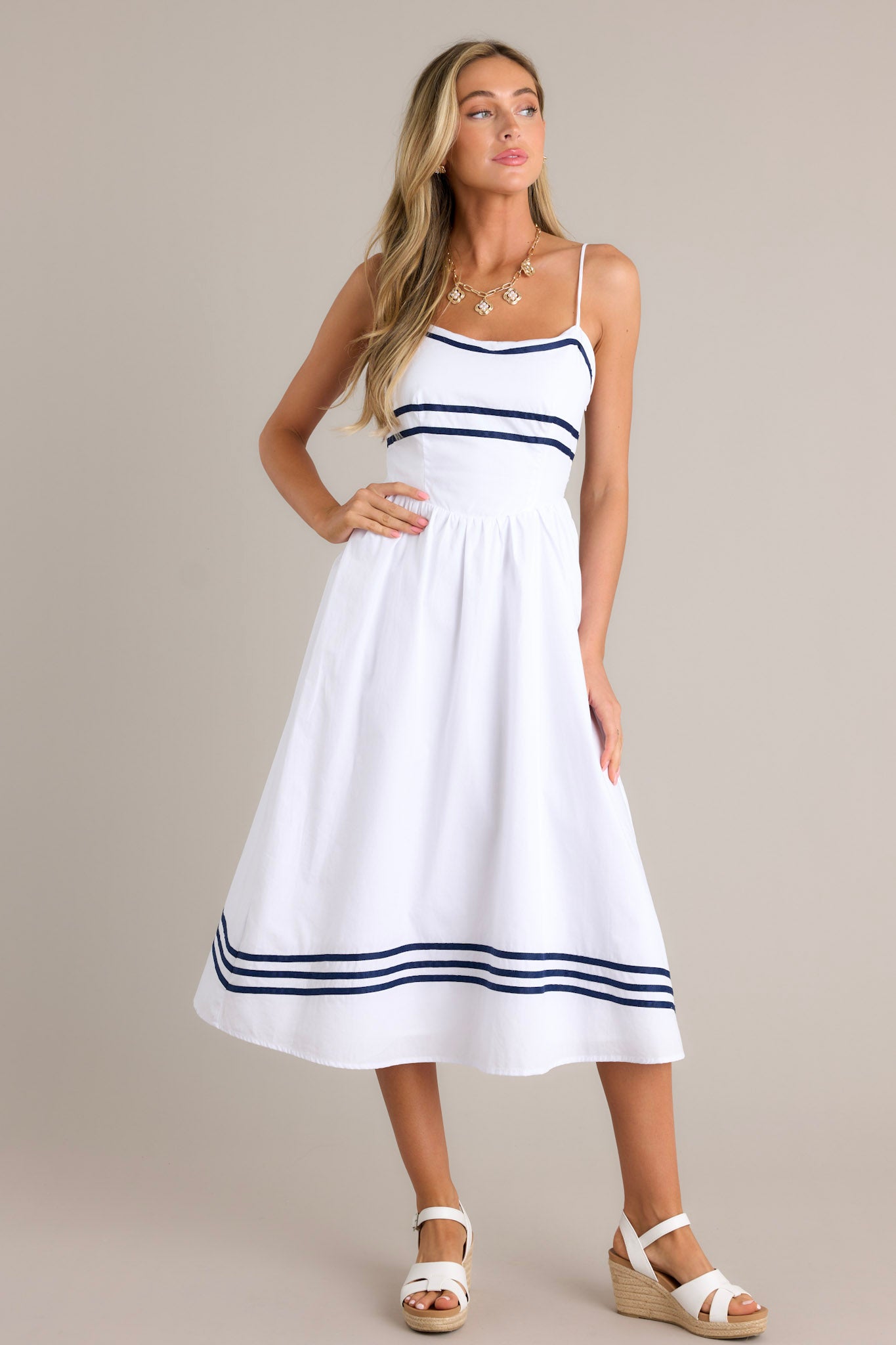 Front angled view of a white midi dress featuring a rounded neckline, thin adjustable straps, a smocked back insert, a discrete side zipper, a dropped waist, navy stripe detailing, and a flowing silhouette