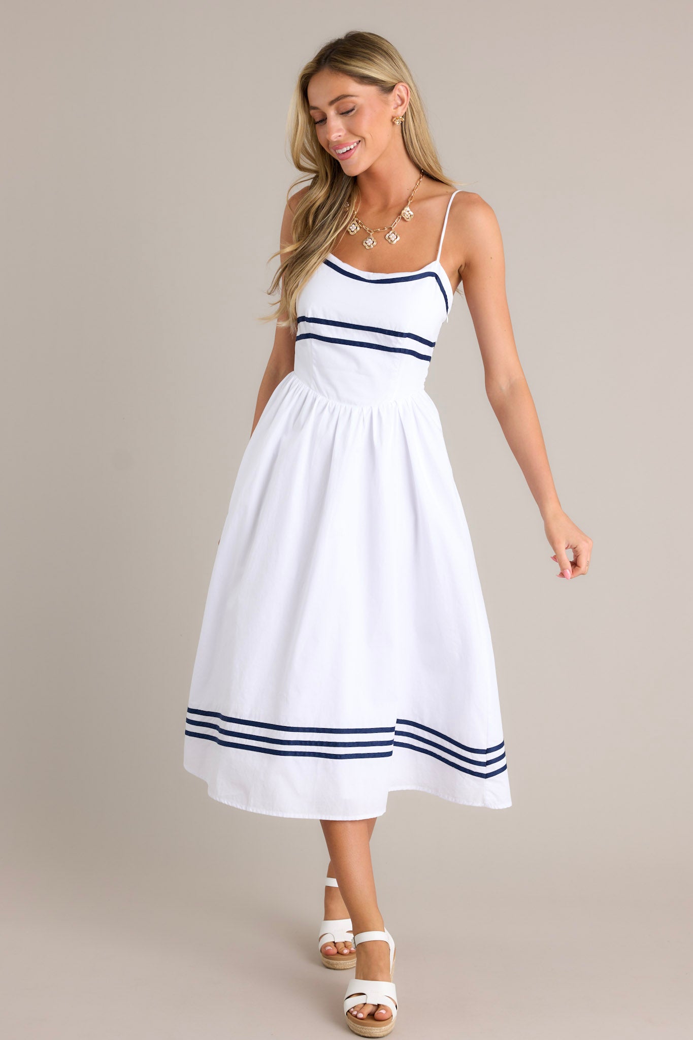 Action shot of a white midi dress displaying the fit and movement, highlighting the rounded neckline, thin adjustable straps, smocked back insert, dropped waist, navy stripe detailing, and flowing silhouette.