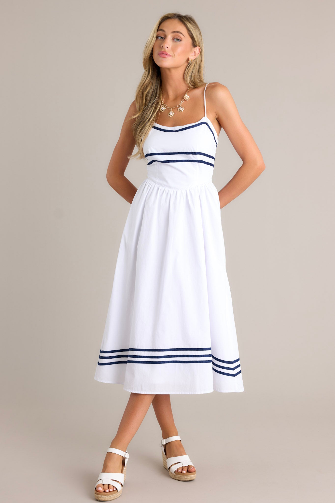 This white midi dress features a rounded neckline, thin adjustable straps, a smocked back insert, a discrete side zipper, a dropped waist, navy stripe detailing, and a flowing silhouette.