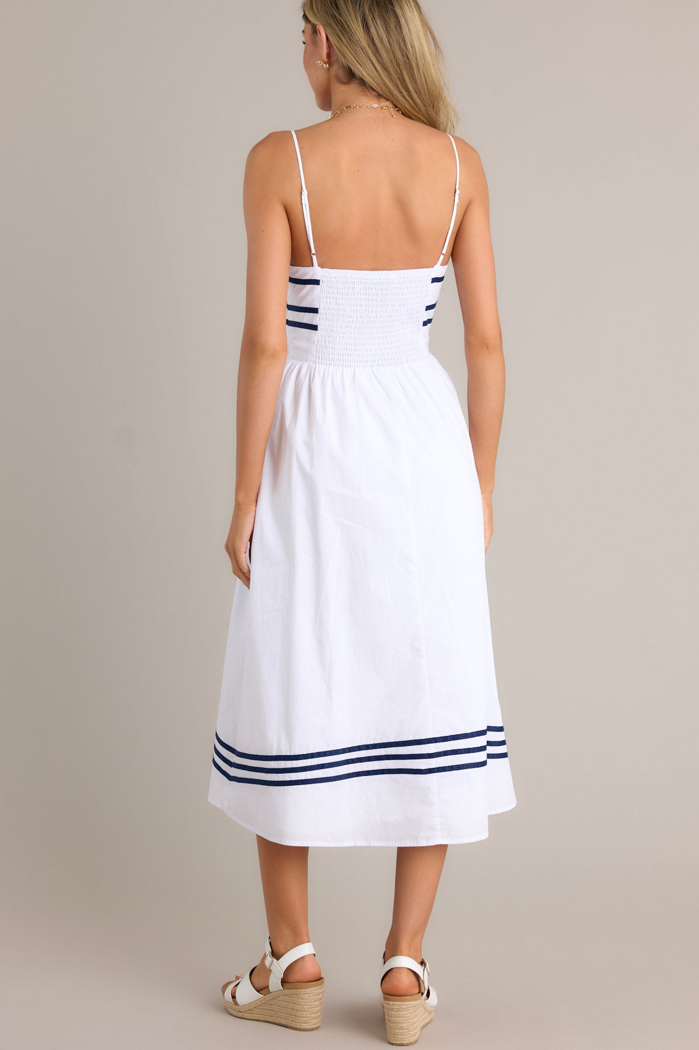 Back view of a white midi dress highlighting the smocked back insert, thin adjustable straps, and navy stripe detailing.