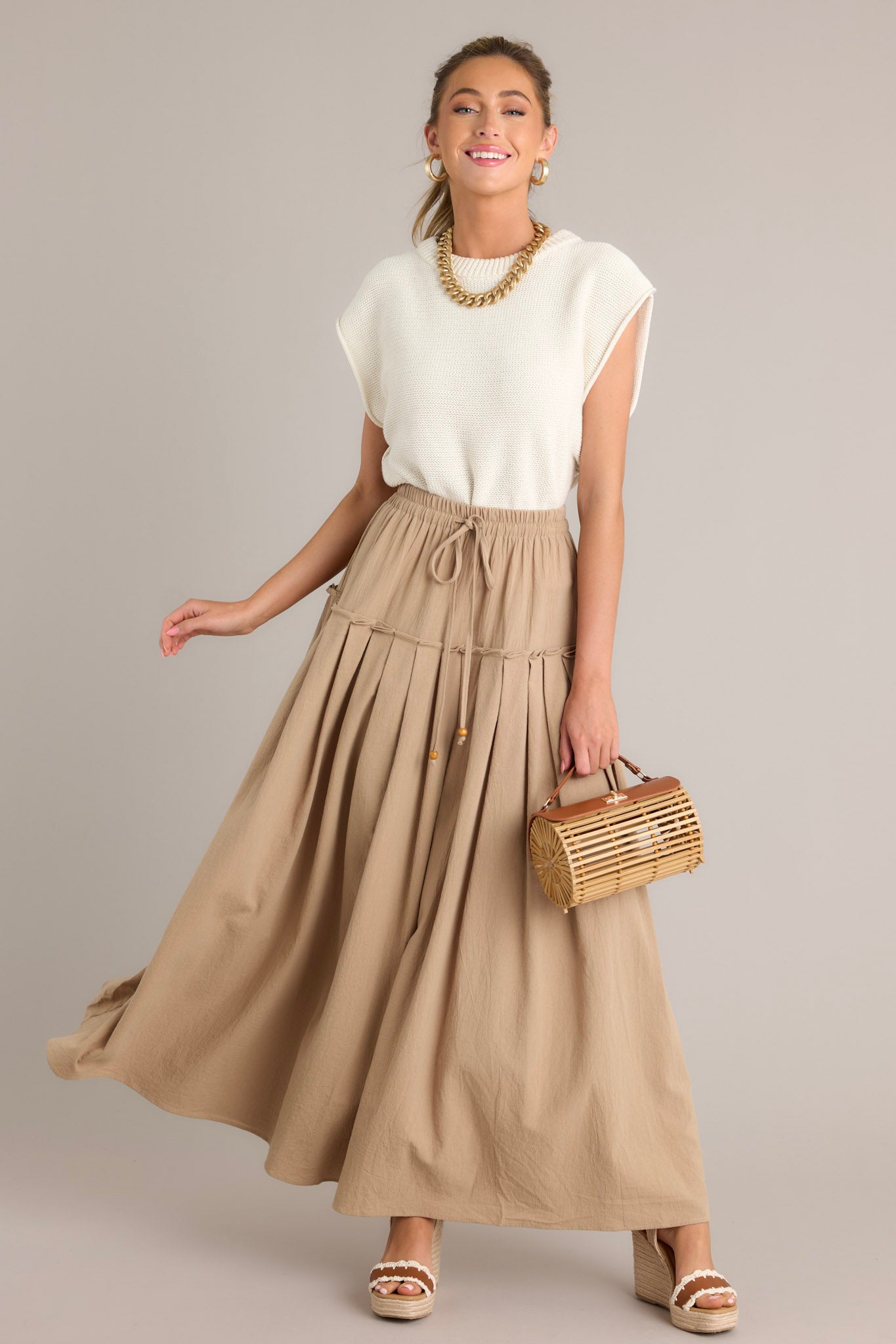 This beige maxi skirt features a high waisted design, an elastic waistband, a self-tie drawstring, a single tier, subtle pleats, and a flowing silhouette.