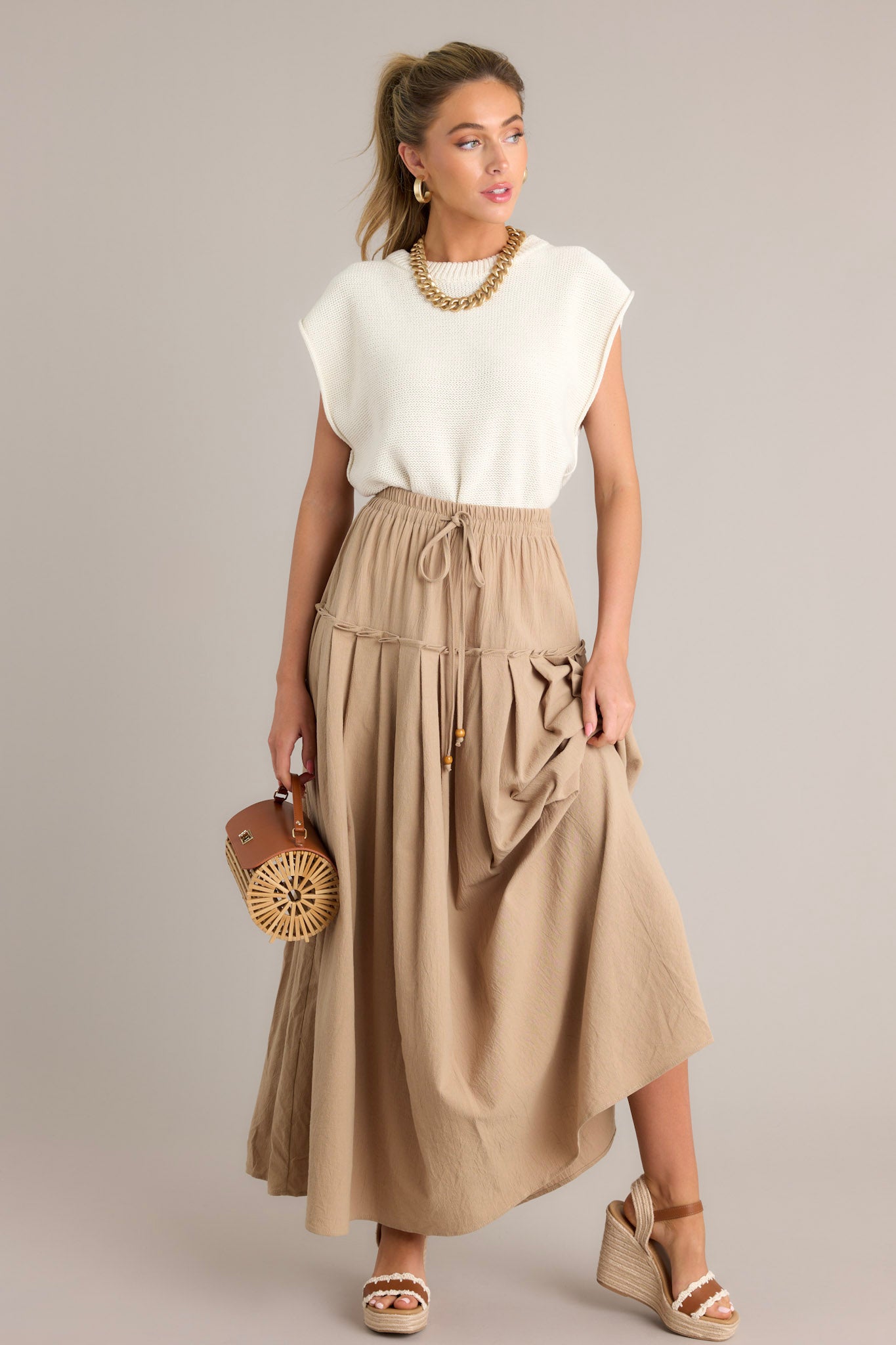 Front angled view of a beige maxi skirt featuring a high waisted design, an elastic waistband, a self-tie drawstring, a single tier, subtle pleats, and a flowing silhouette