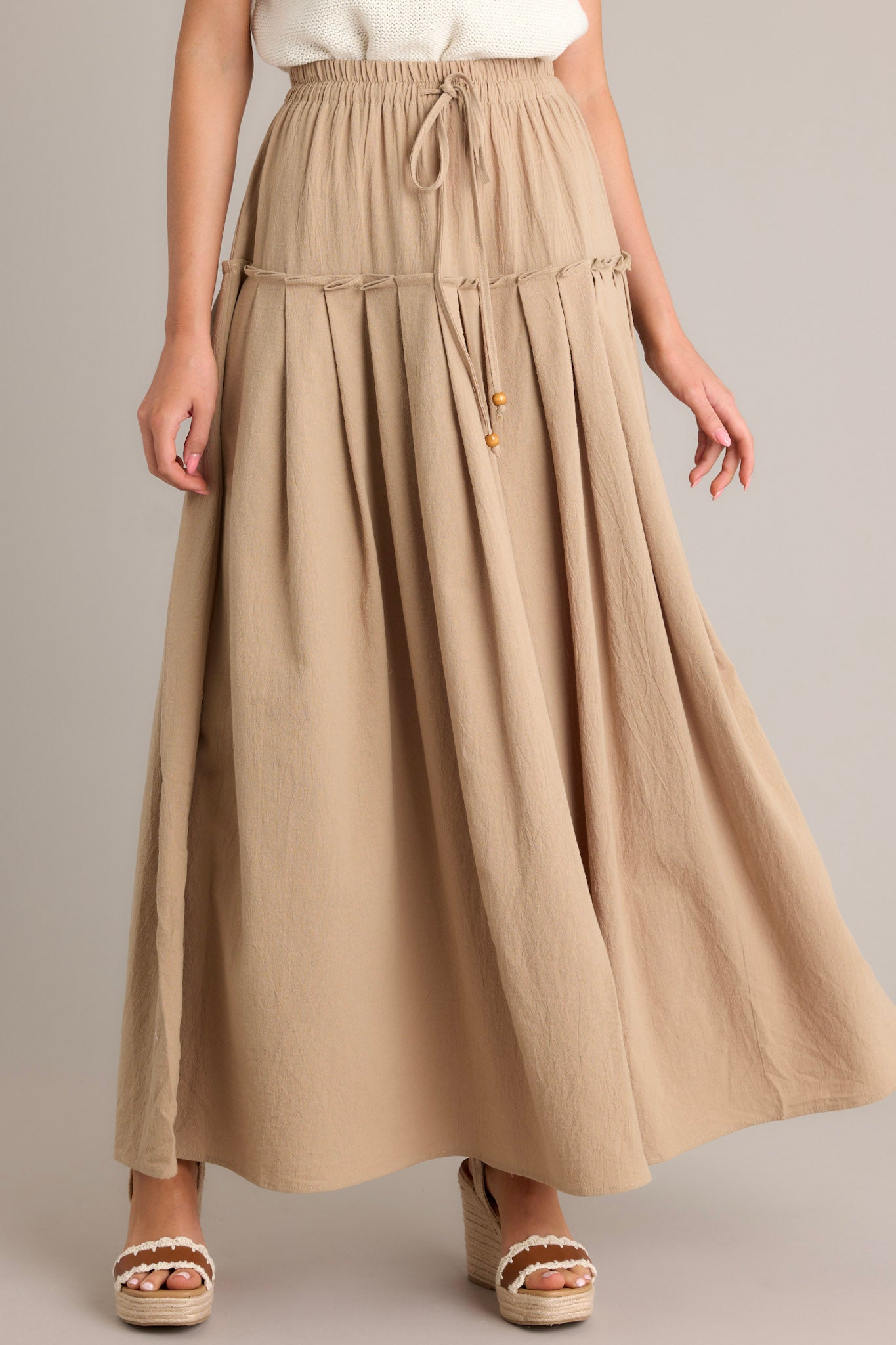 Front view of a beige maxi skirt featuring a high waisted design, an elastic waistband, a self-tie drawstring, a single tier, subtle pleats, and a flowing silhouette.