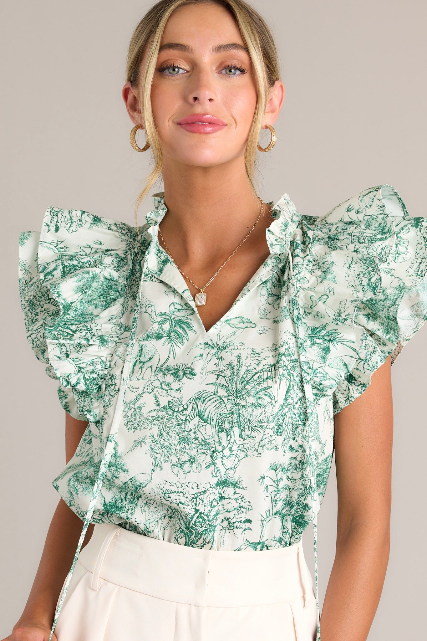 Front view of a green top featuring a ruffled v-neckline with a self-tie closure, a lightweight material, ruffle sleeves, and a toile print.